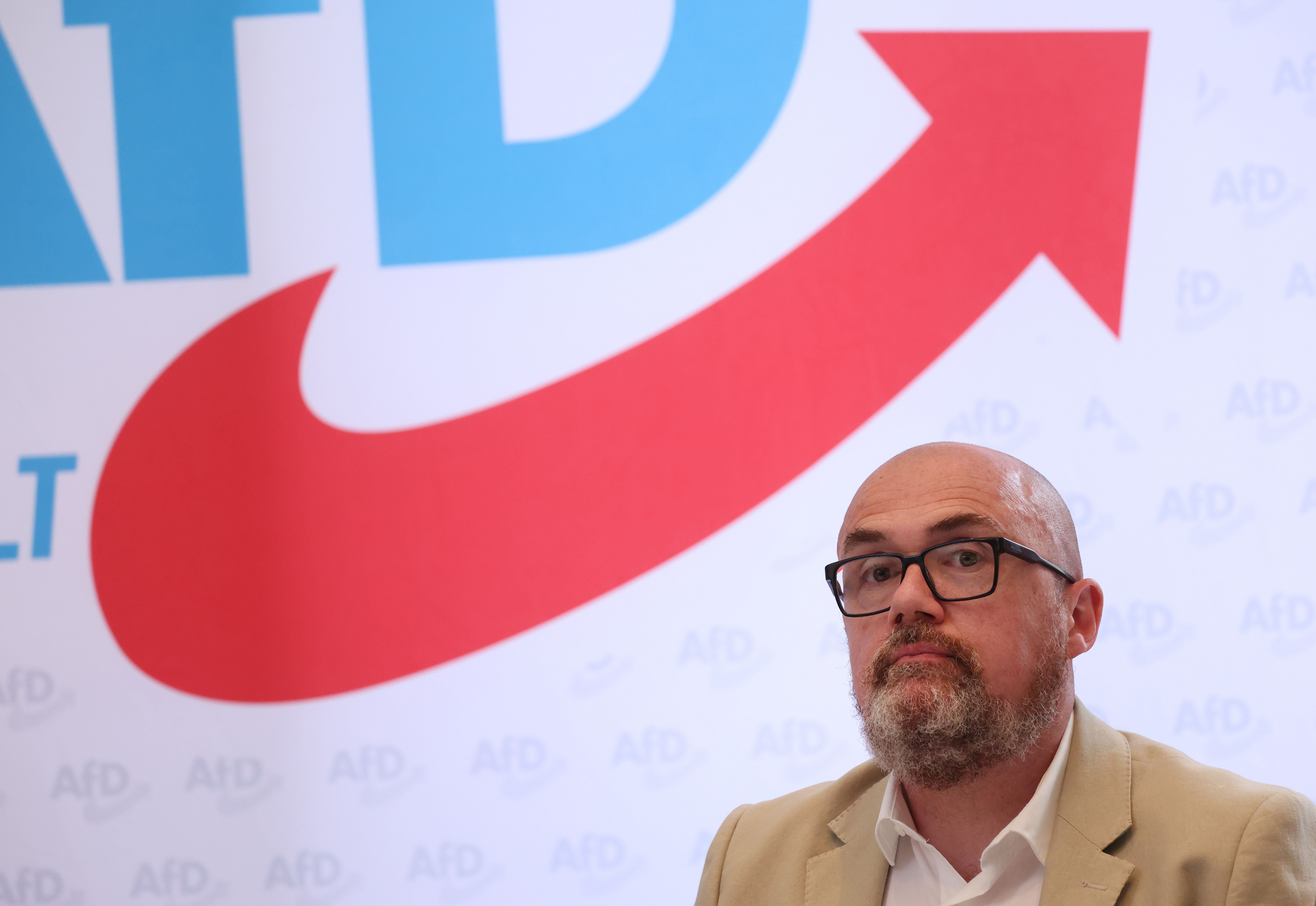 Ein Mann mit Brille und Bart sitzt vor einem AfD-Logo mit großem roten Pfeil.