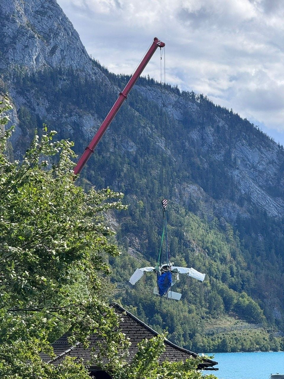 Ein Kran hebt ein Flugzeugwrack über ein Hausdach vor einer Berglandschaft.
