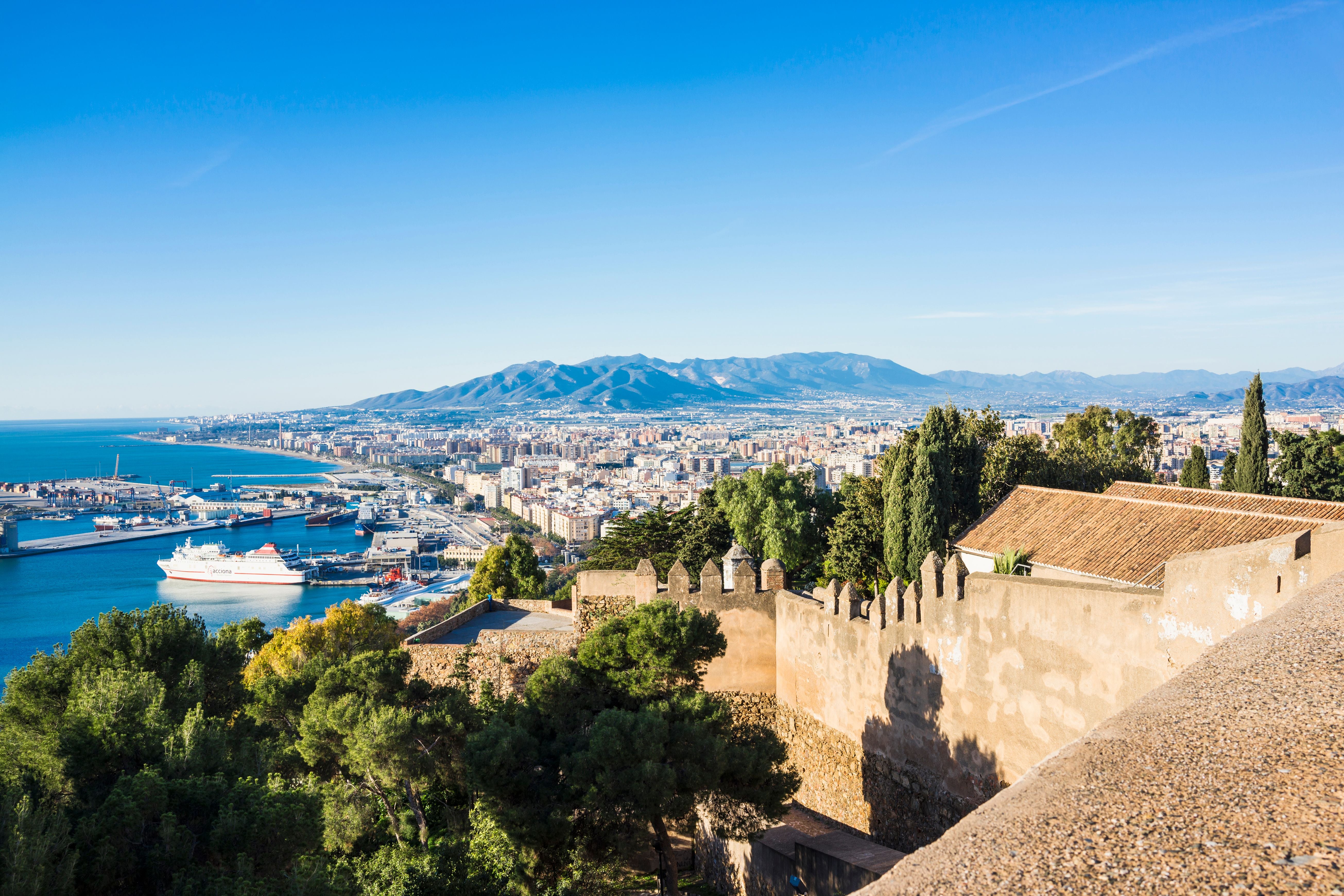 Blick vom Castillo de Gibralfaro auf den Hafen von Málaga und die Stadt mit Bergen im Hintergrund.