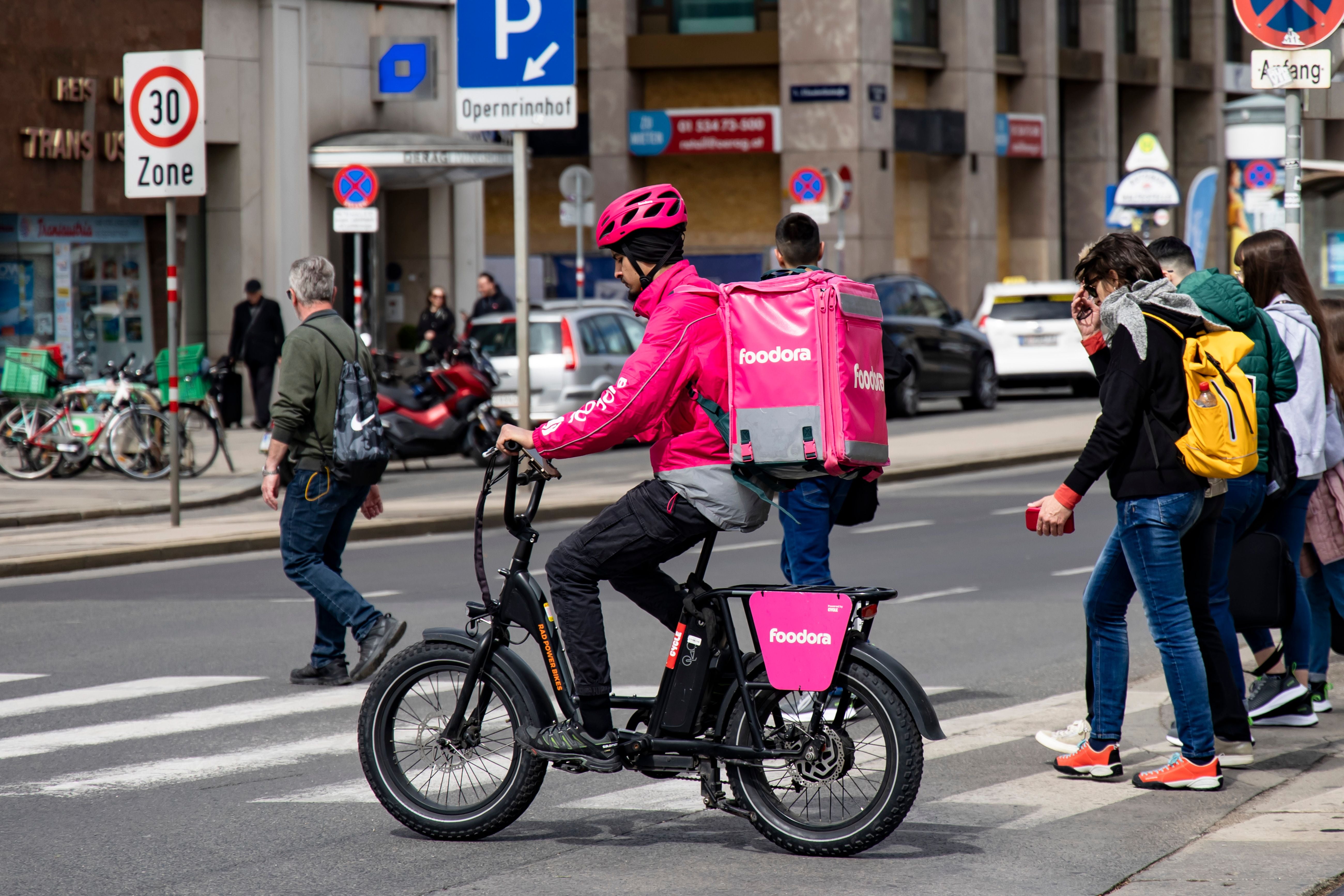 Ein Foodora-Lieferant fährt mit dem Fahrrad durch eine Innenstadt in Wien, umgeben von Passanten.