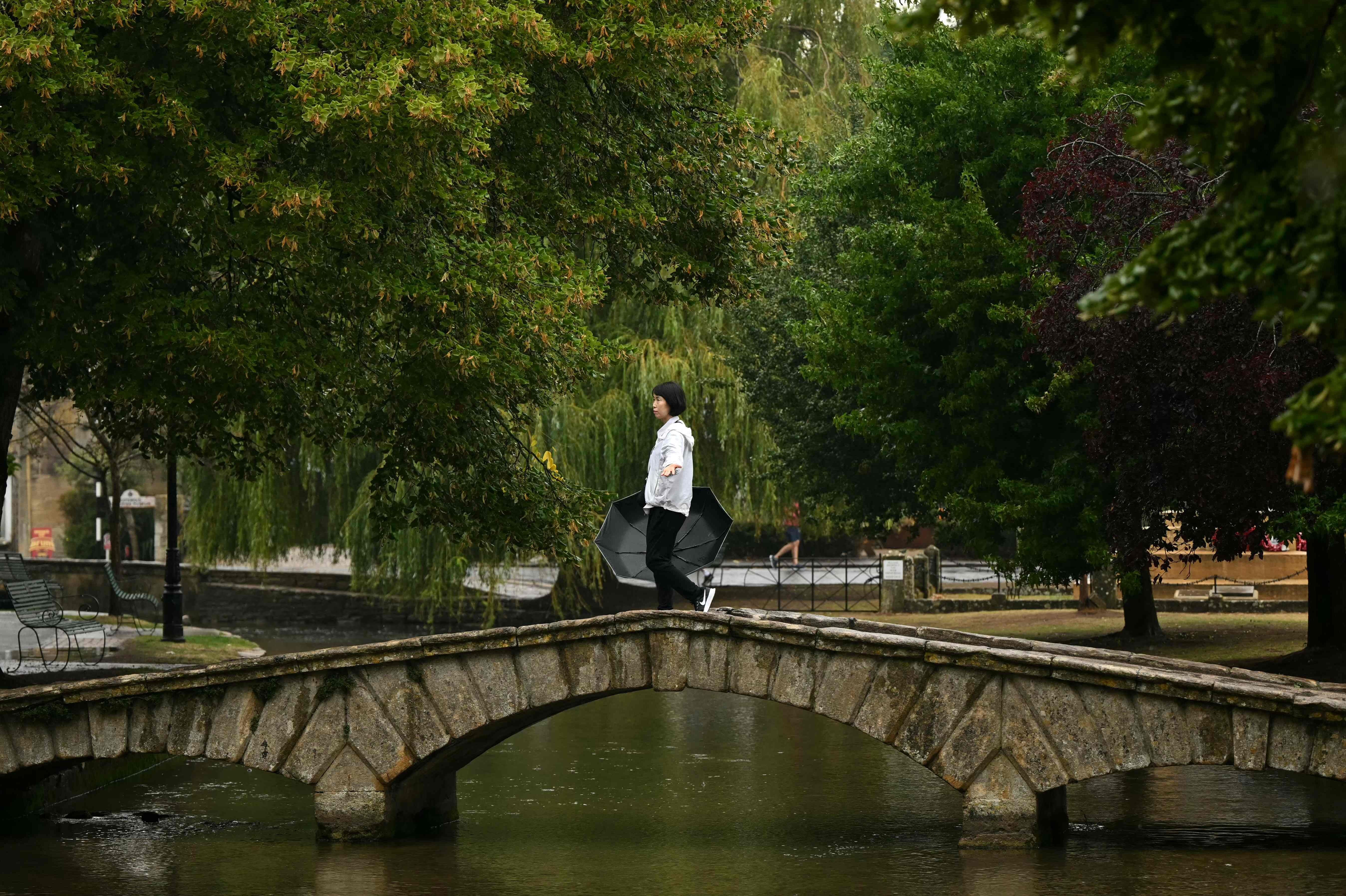 Eine Person mit Regenschirm geht über eine steinerne Brücke inmitten grüner Bäume.