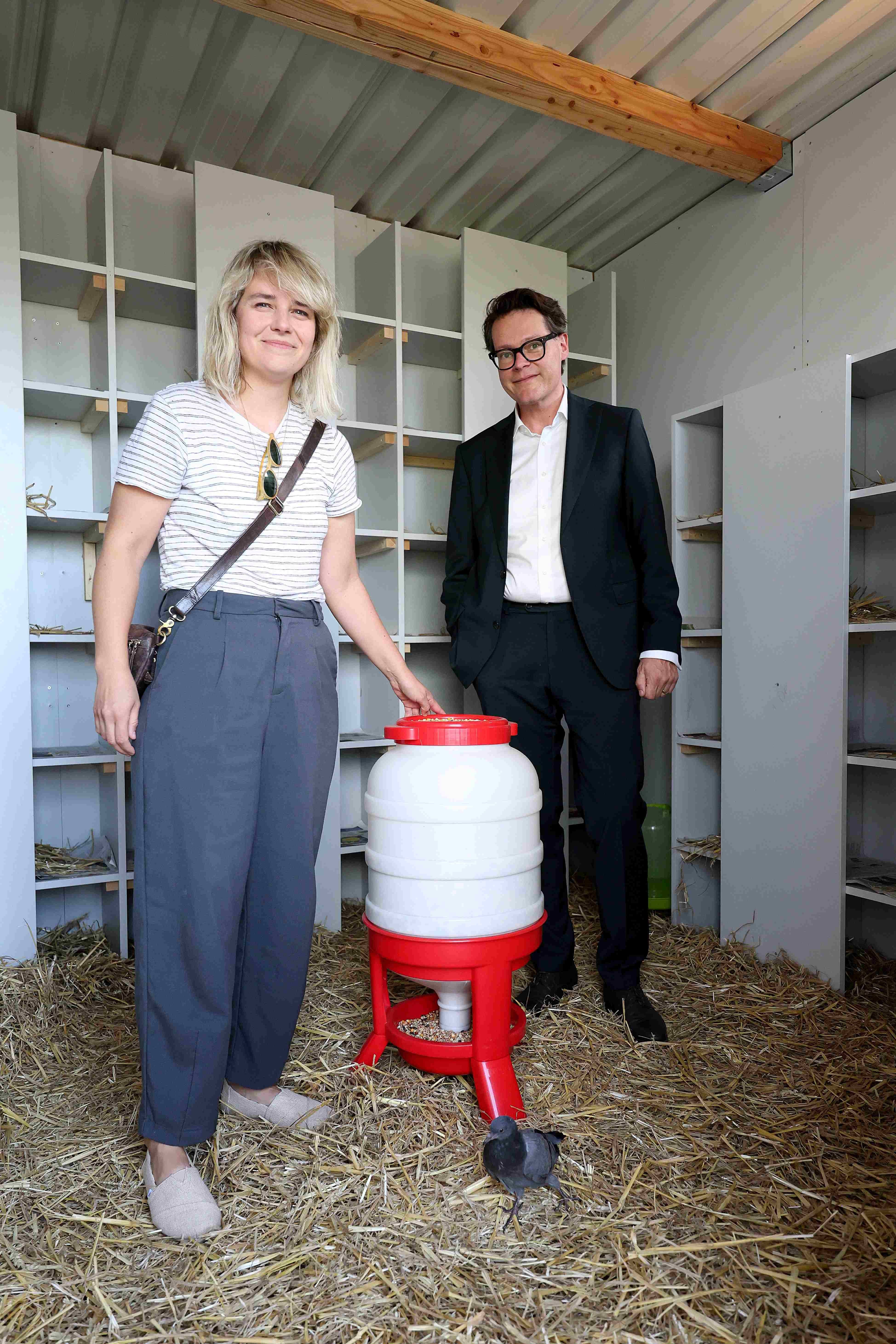 Eine Frau und ein Mann stehen in einem Taubenschlag neben einem Futterspender und einer Taube.