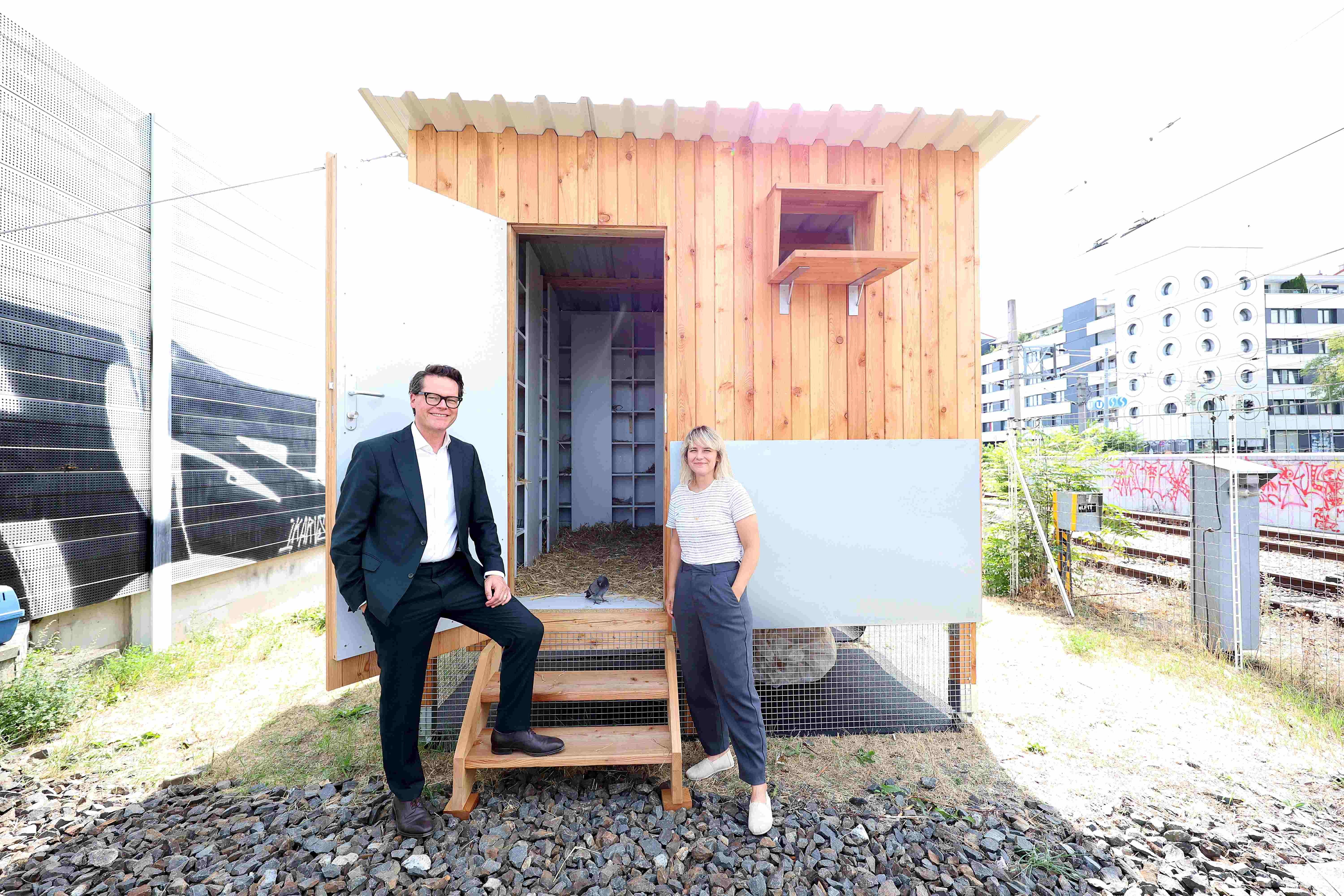 Zwei Personen stehen vor einem mobilen Taubenschlag aus Holz in einem urbanen Umfeld.