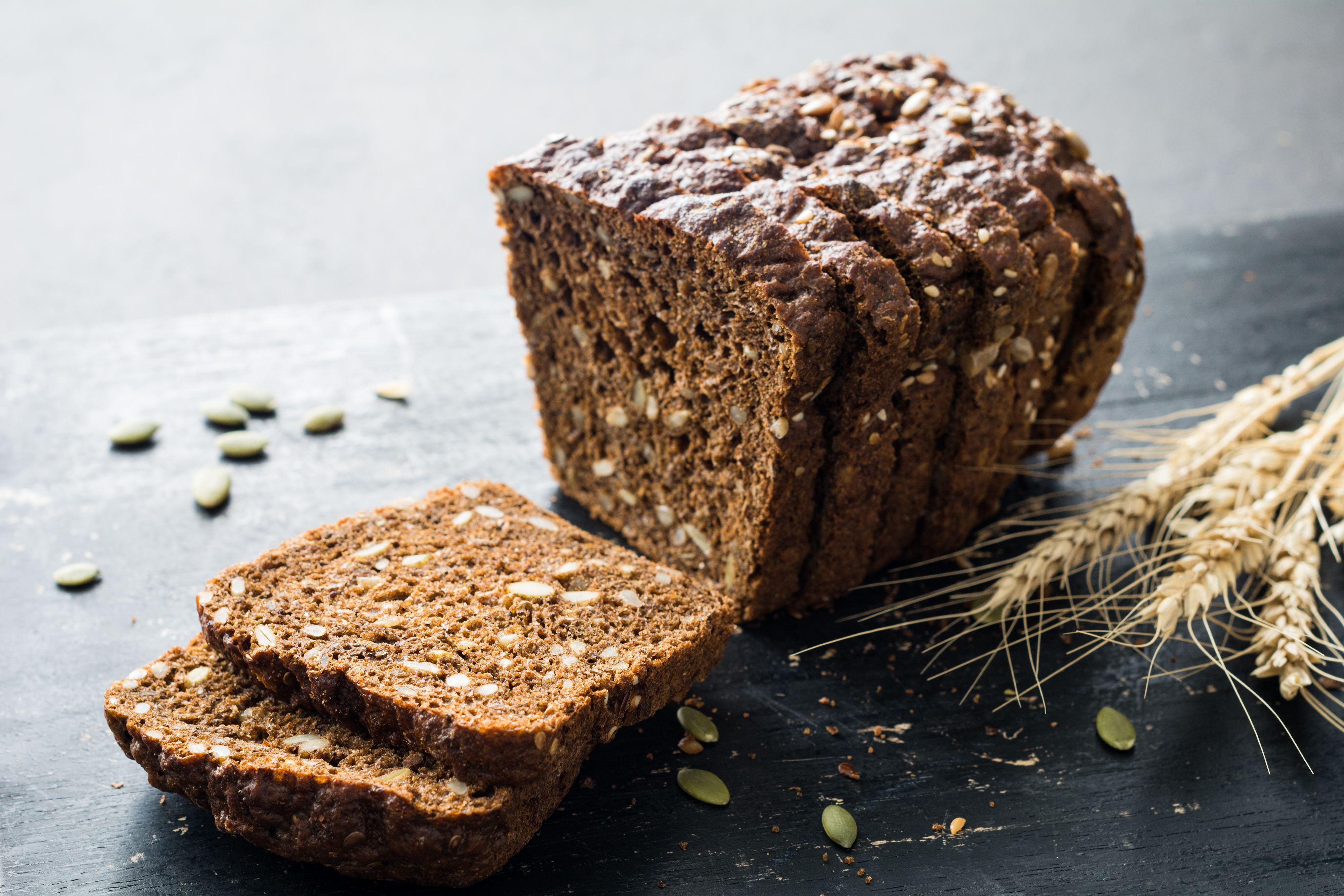 Frisches Vollkorn-Roggenbrot mit Körnern auf einem schwarzen Brett, daneben Getreideähren.