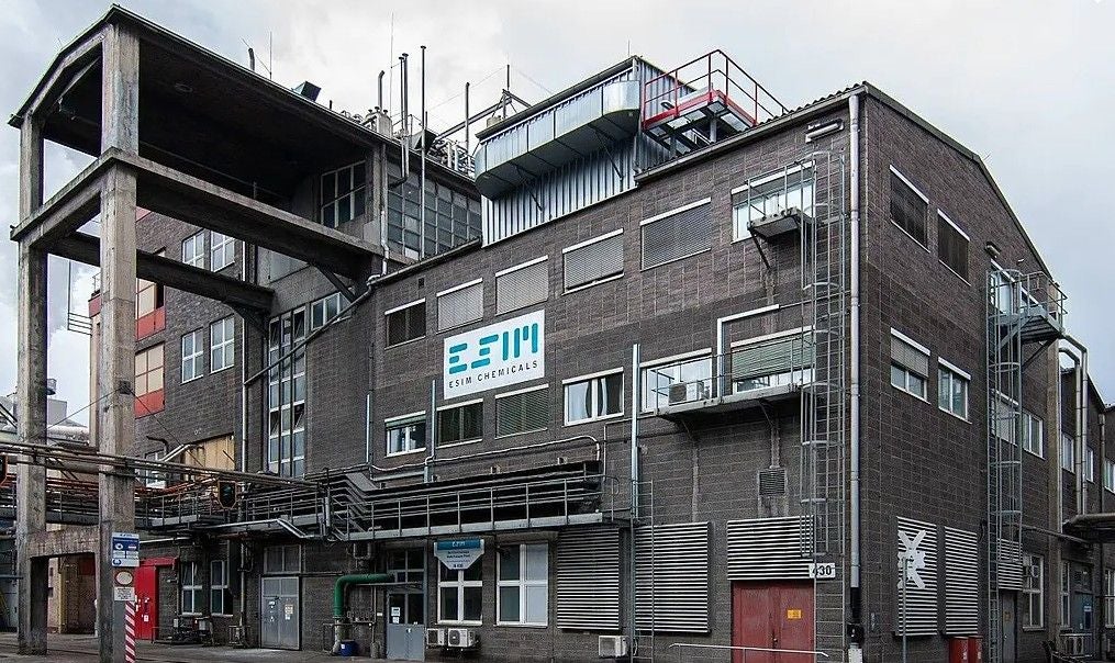 Industriegebäude mit vielen Rohren; Schild „ESIM CHEMICALS“ an der Fassade.