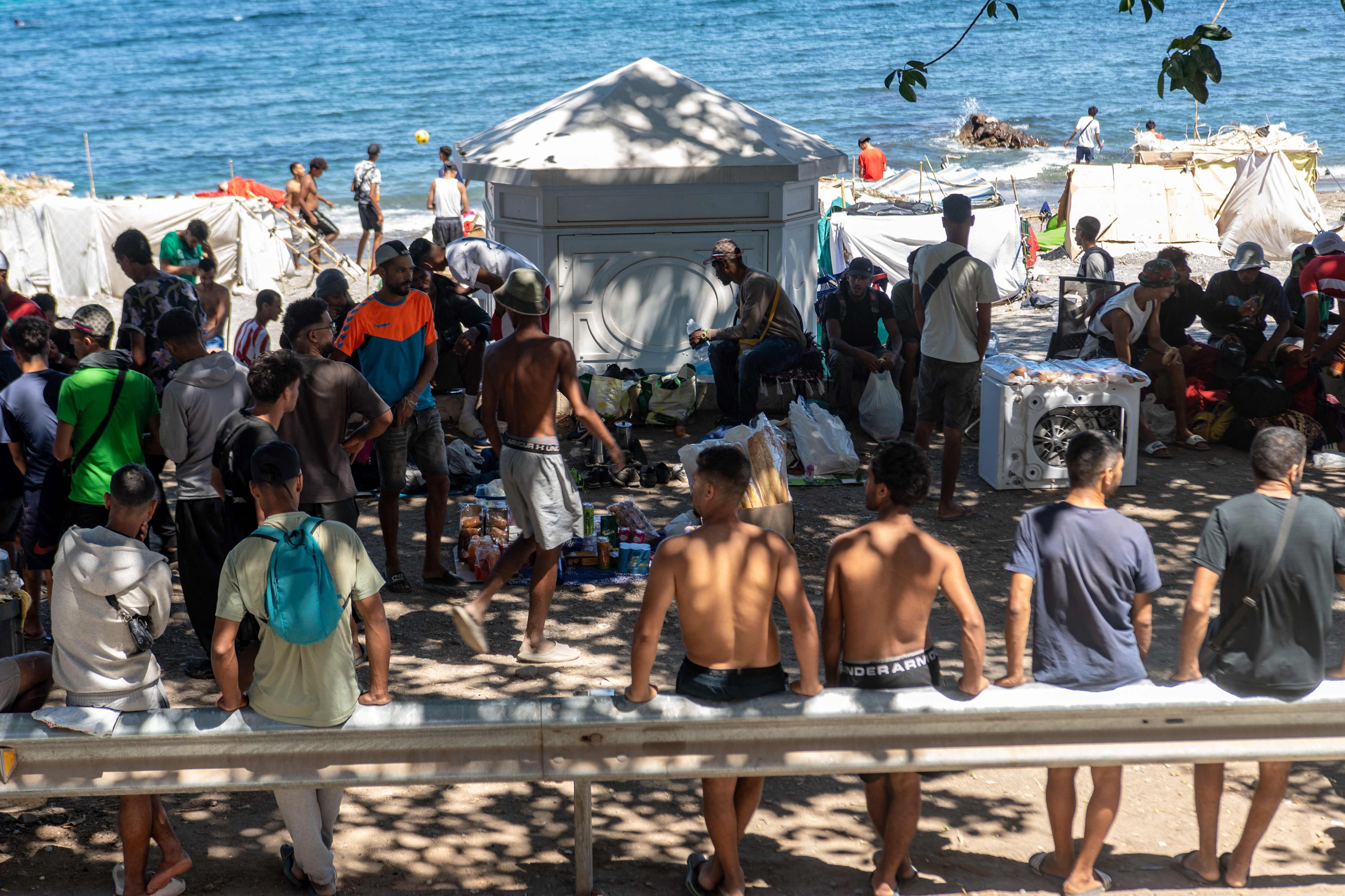 Eine große Gruppe Migranten versammelt sich neben Zelten am Strand, im Hintergrund ist das Meer zu sehen.