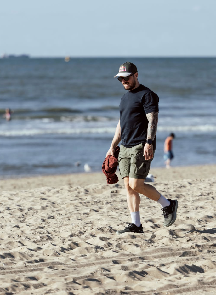 Ein Mann mit Sonnenbrille und Mütze spaziert entspannt am Sandstrand entlang.