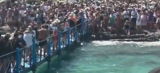 Viele Menschen stehen dicht gedrängt auf einer Brücke und am Ufer über türkisfarbenem Wasser.