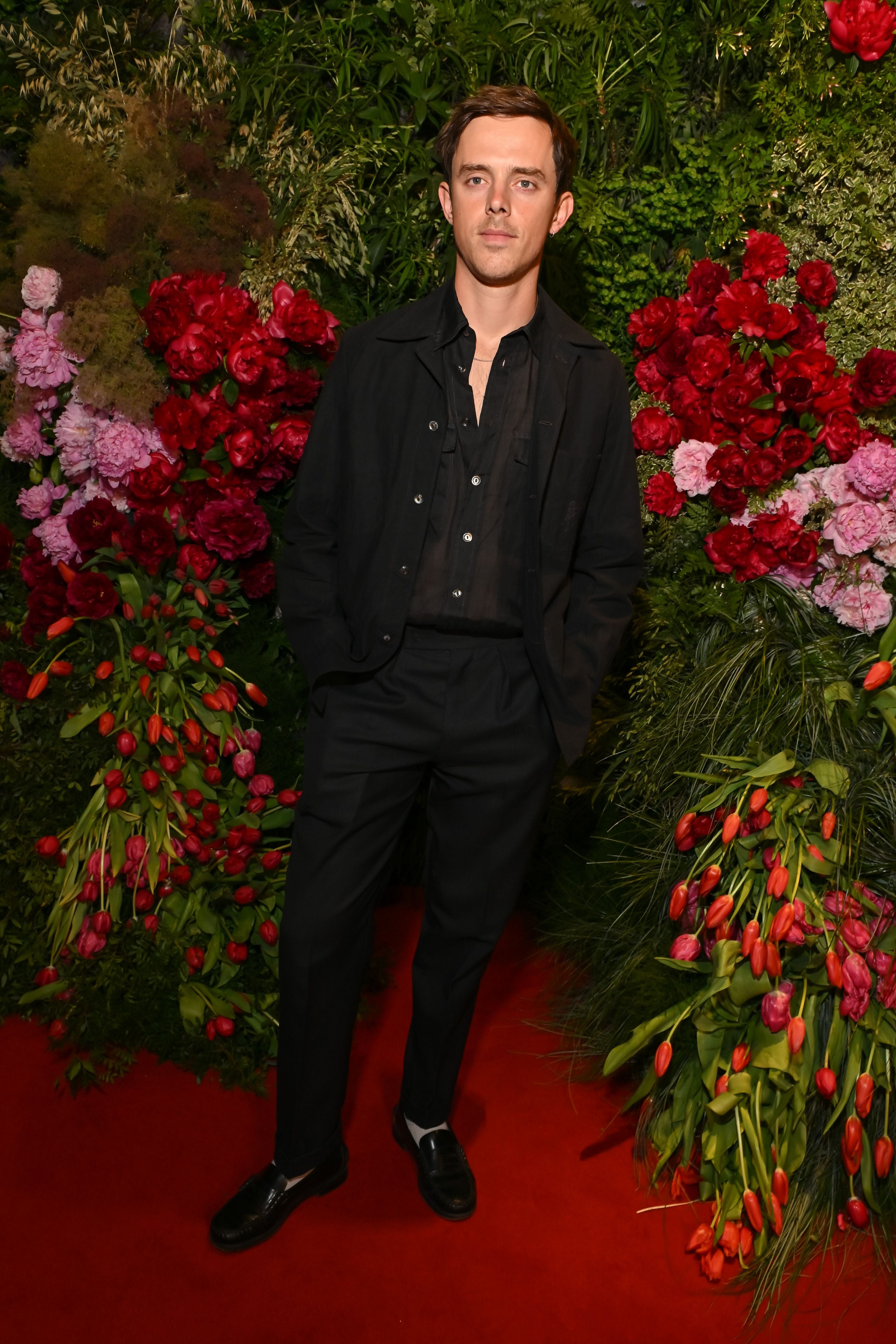 Ein Mann in dunkler Kleidung steht auf rotem Teppich vor Blumenwand mit roten und rosa Blüten.