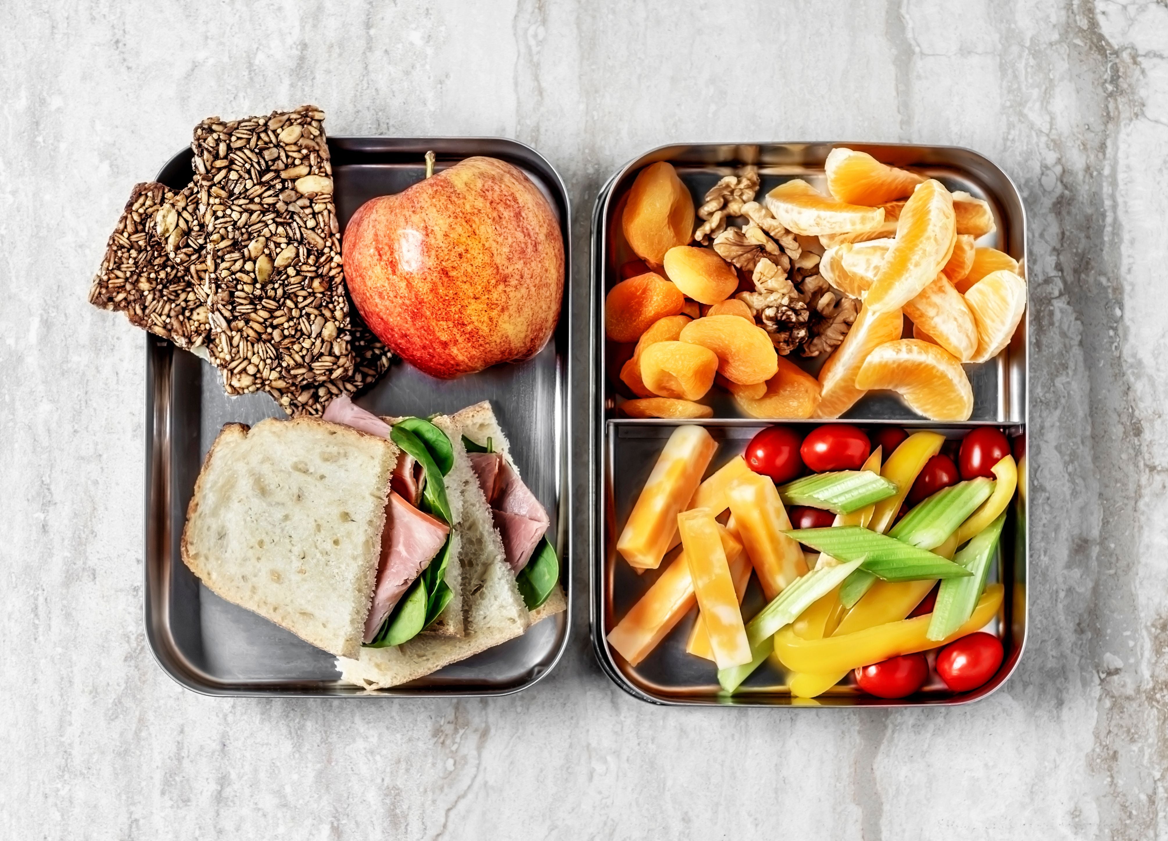 Zwei Brotdosen mit Sandwich, Apfel, Saaten-Crackern, Nüssen, Trockenfrüchten und Gemüse.