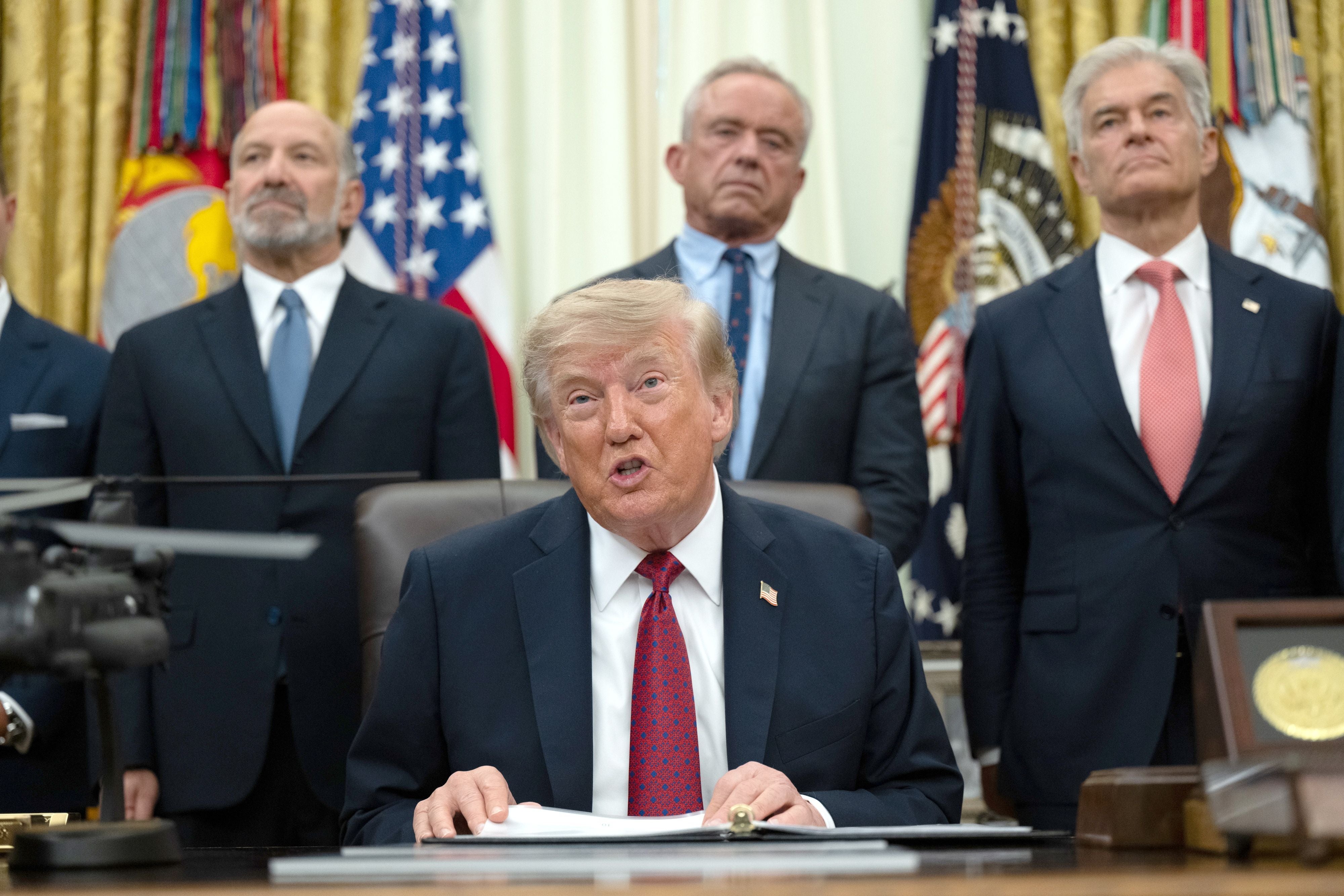Donald Trump sitzt am Schreibtisch im Oval Office, drei Männer stehen hinter ihm.