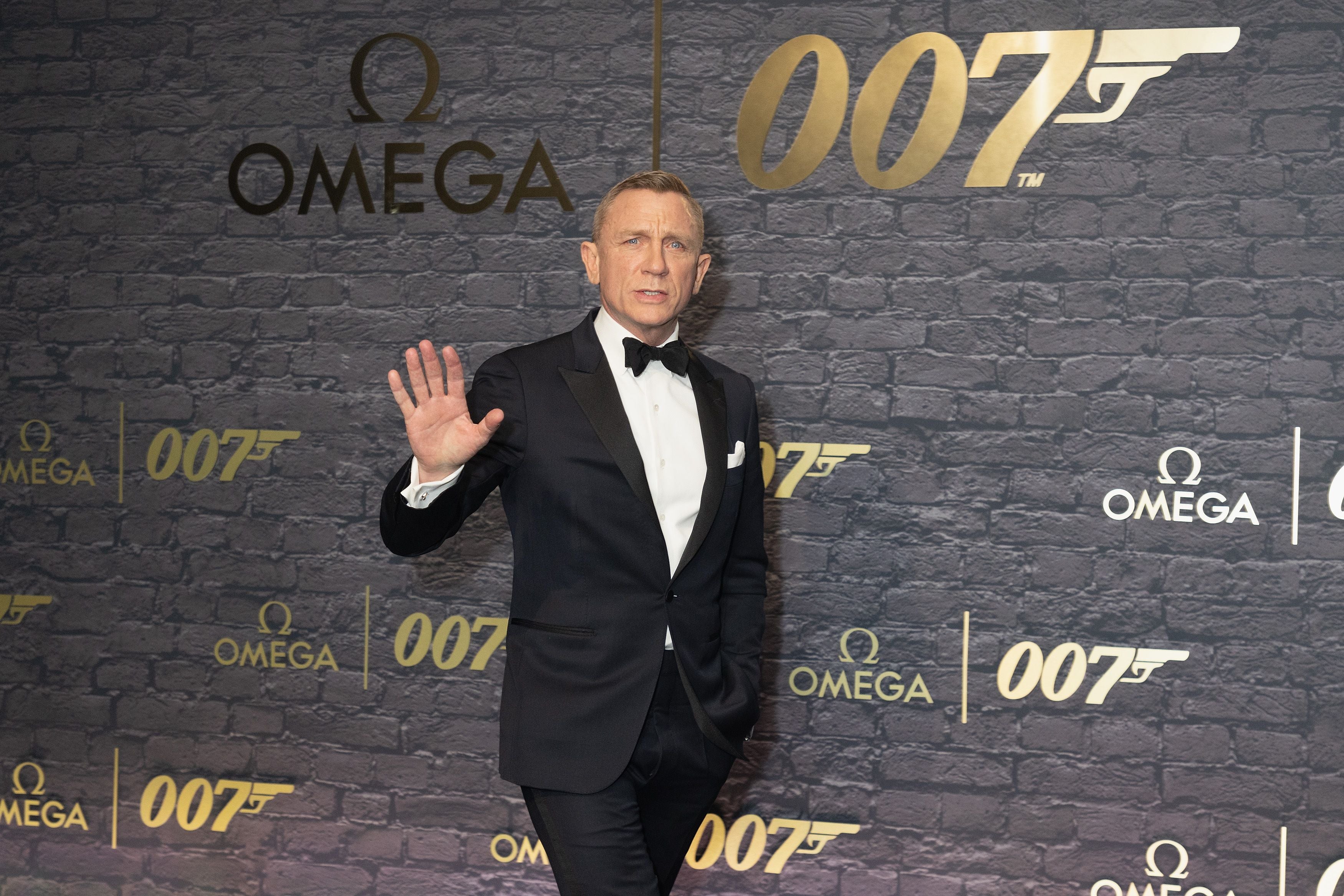 Ein Mann im Smoking winkt vor einer Wand mit Omega- und 007-Logos in London.