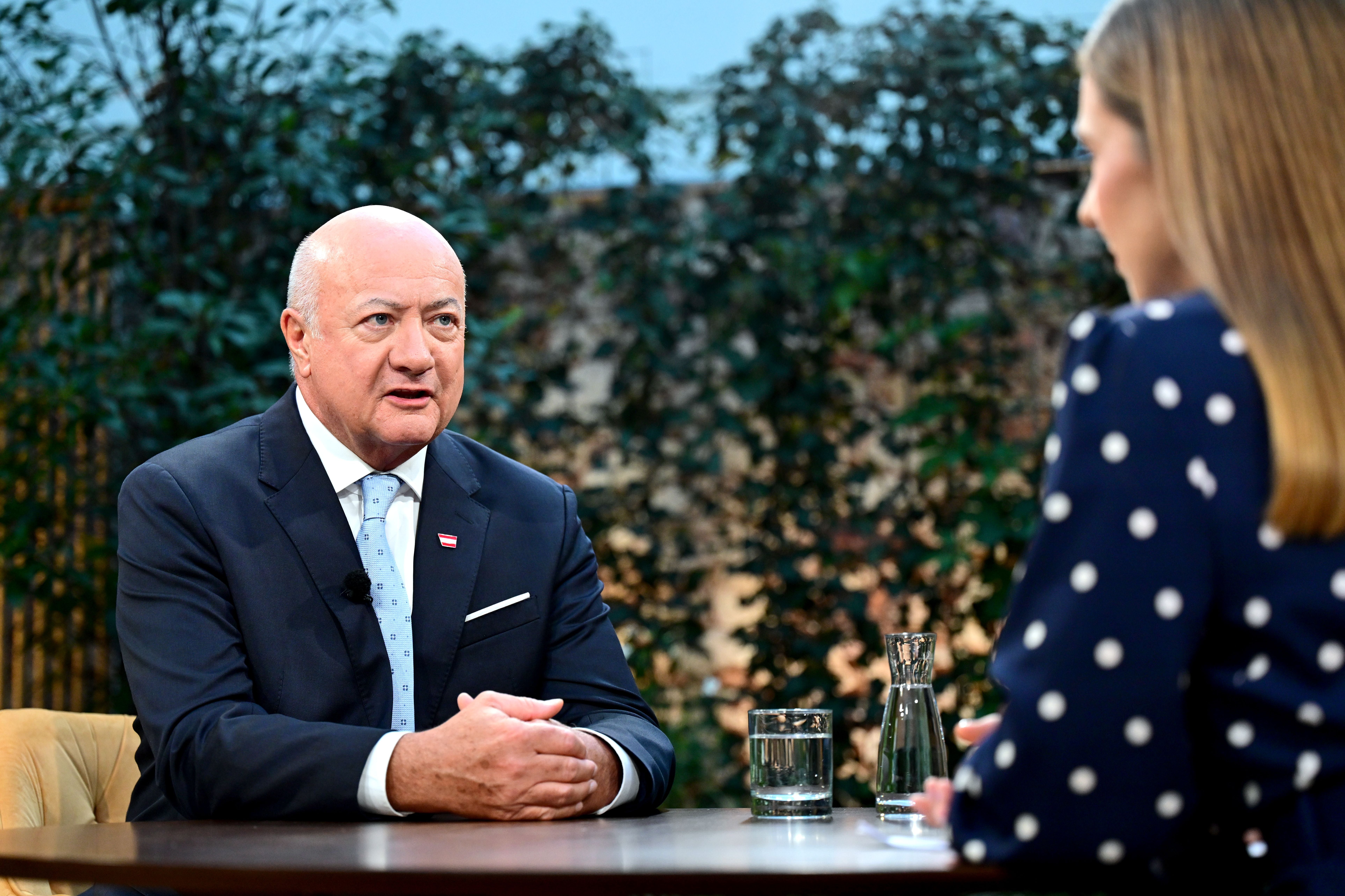 Zwei Personen führen ein Interview an einem Tisch mit Wasserglas und Karaffe, im Hintergrund Pflanzen.