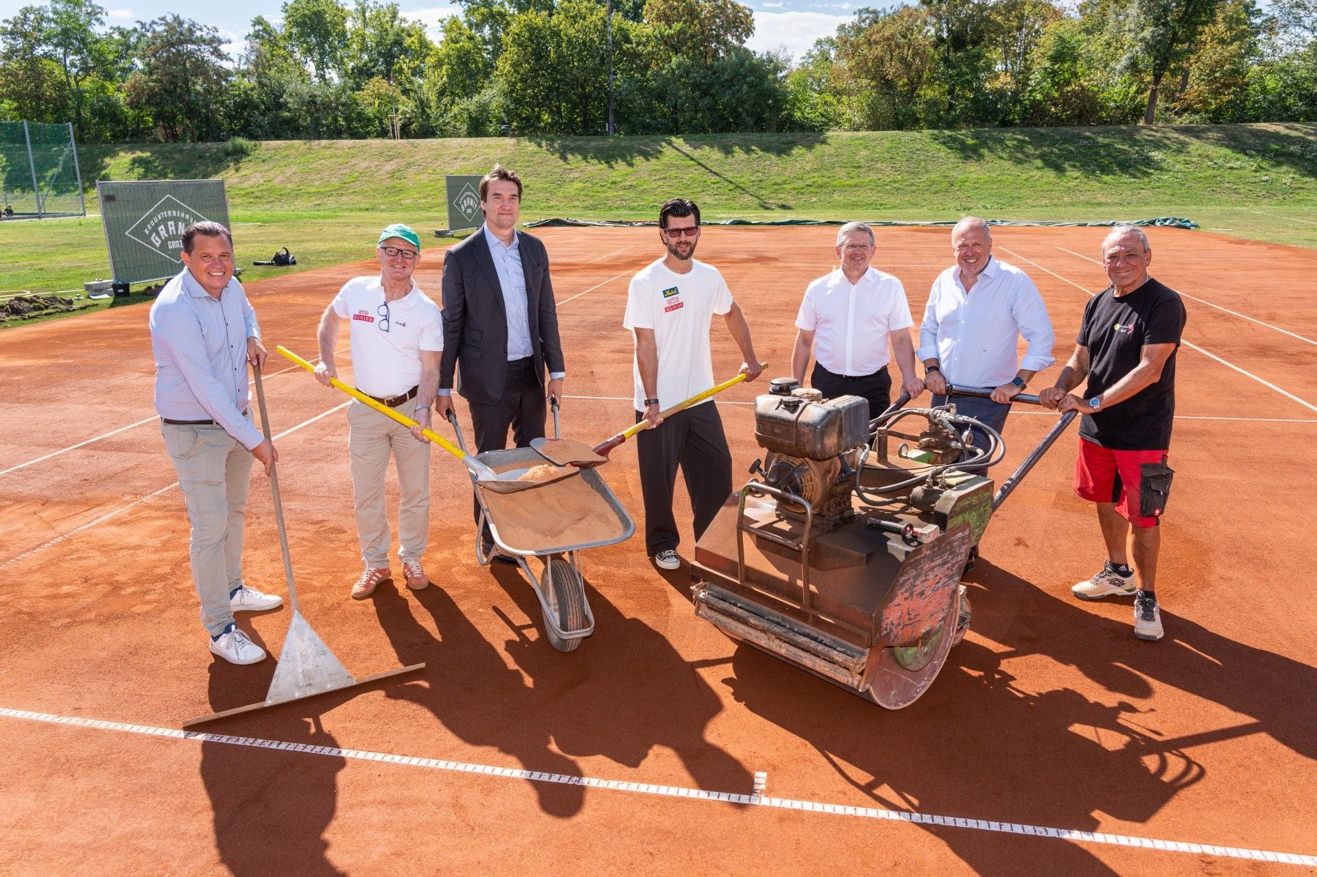 Sieben Männer stehen mit Arbeitsgeräten auf einem frisch hergerichteten Sandtennisplatz.