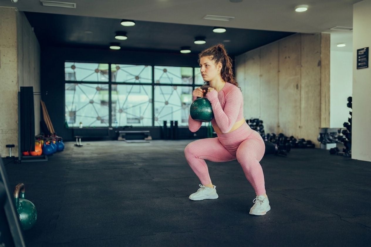 Frau in pinkem Sportoutfit macht Kniebeugen mit einer Kettlebell im Fitnessstudio.