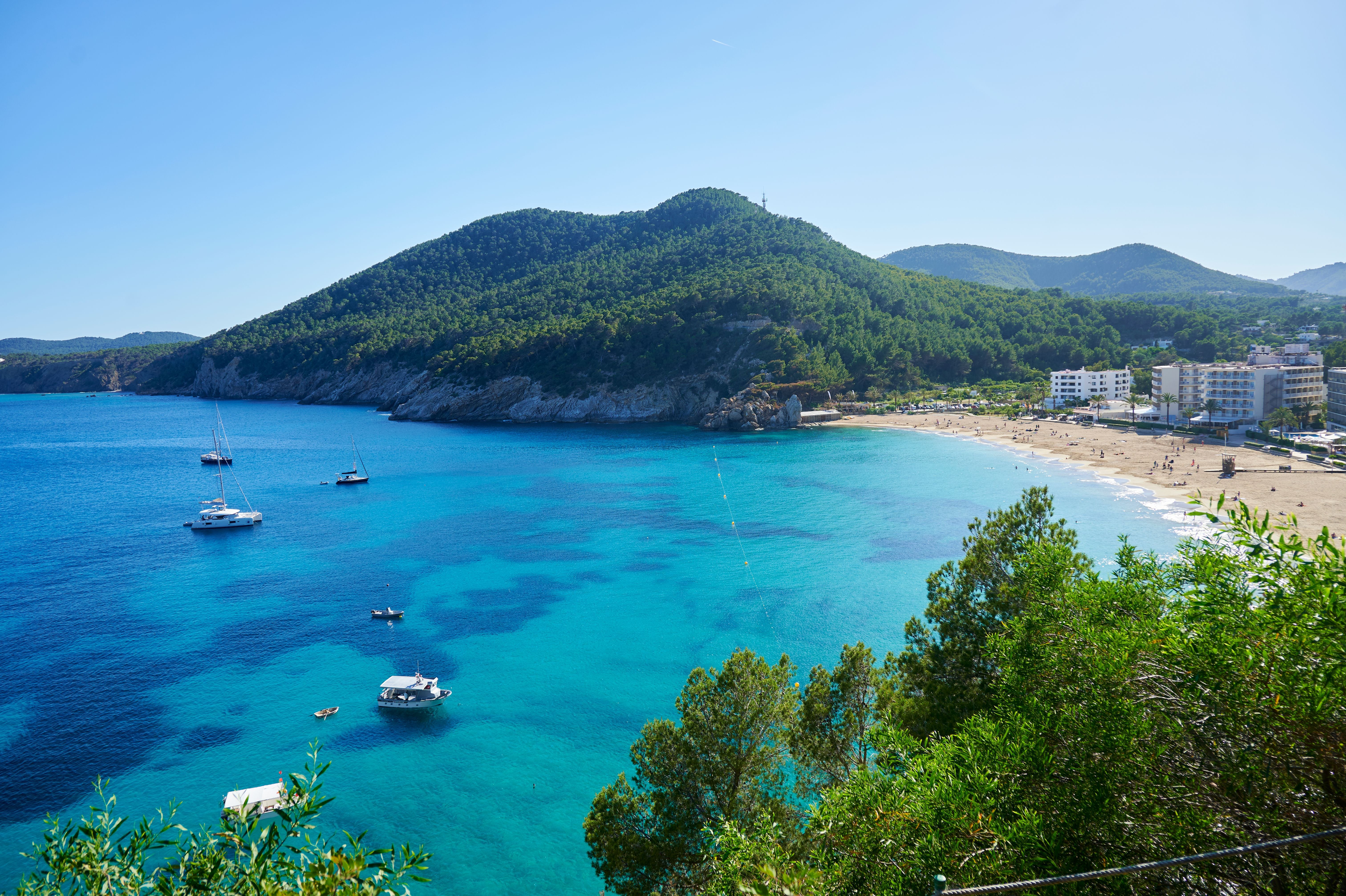 Strand mit türkisblauem Wasser, Segelbooten und bewaldeten Hügeln auf Ibiza, Spanien.