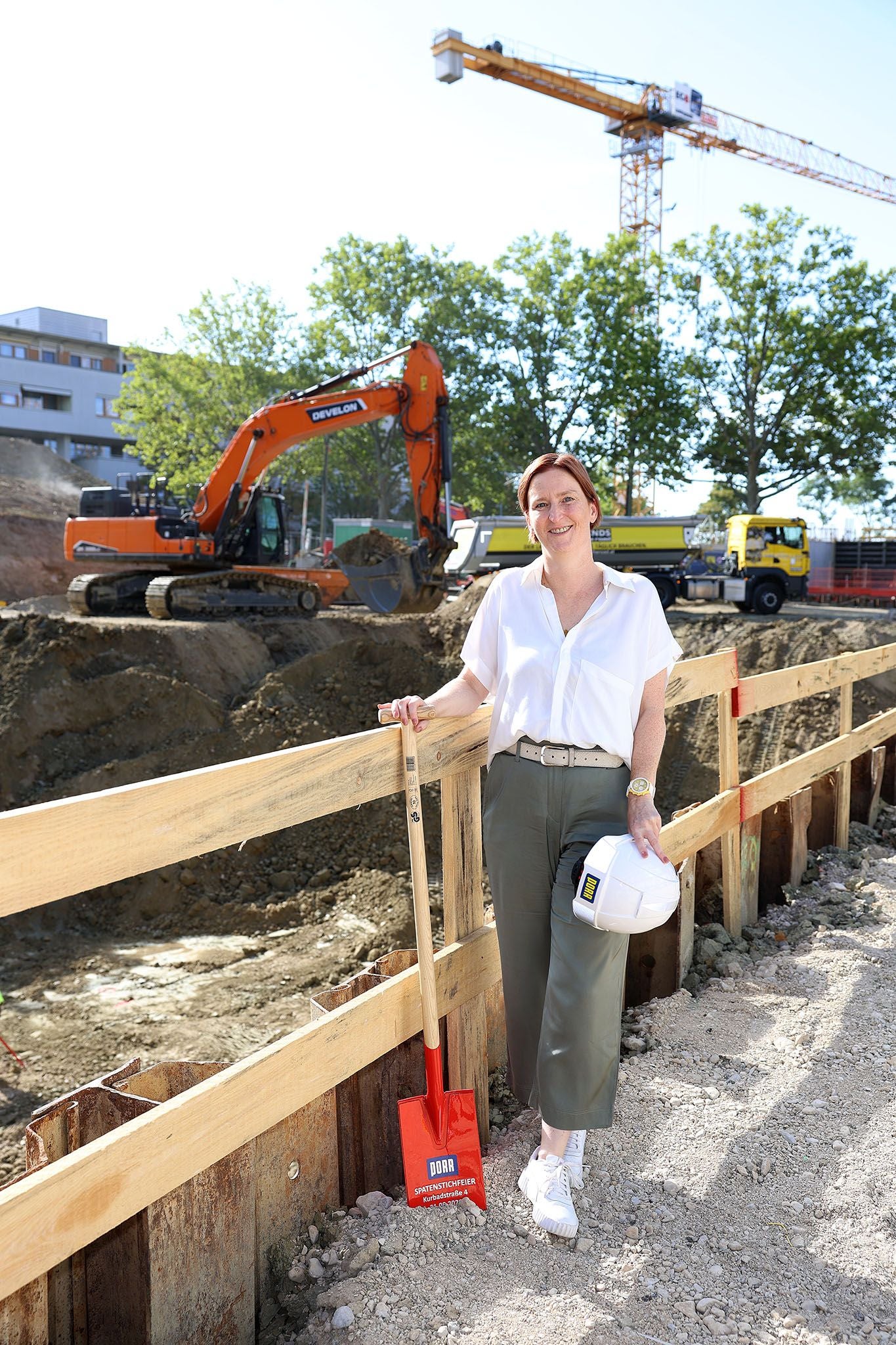 Eine Frau steht mit Spaten und Helm auf einer Baustelle, im Hintergrund Bagger und Kran.