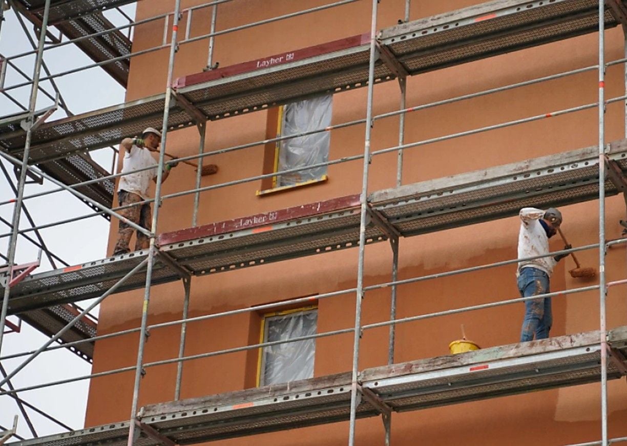 Zwei Bauarbeiter streichen die Fassade eines Wohnblocks auf einem Baugerüst orange an.