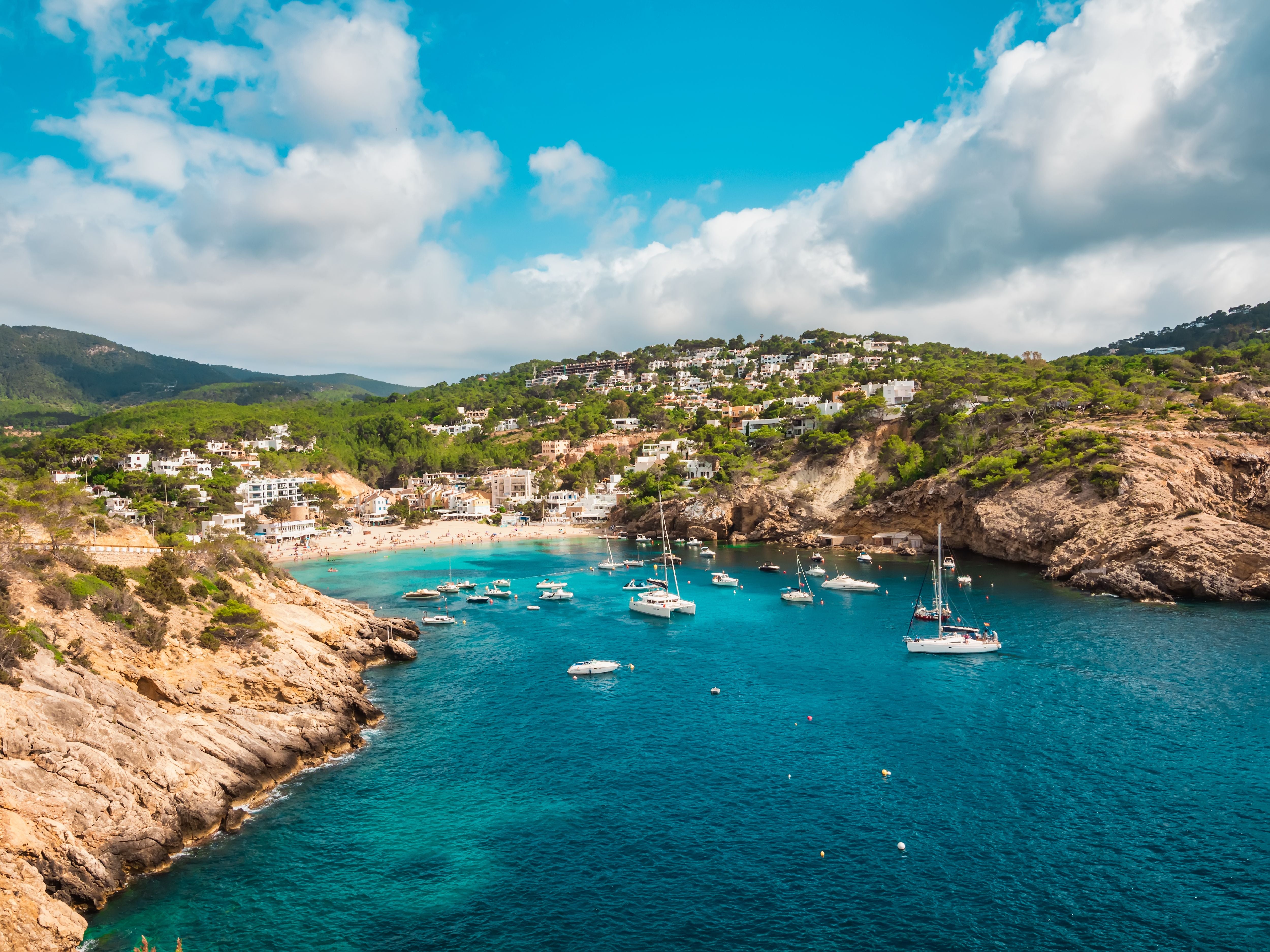 Luftaufnahme der Bucht Cala Vadella auf Ibiza mit Strand, Klippen und Booten im türkisblauen Wasser.
