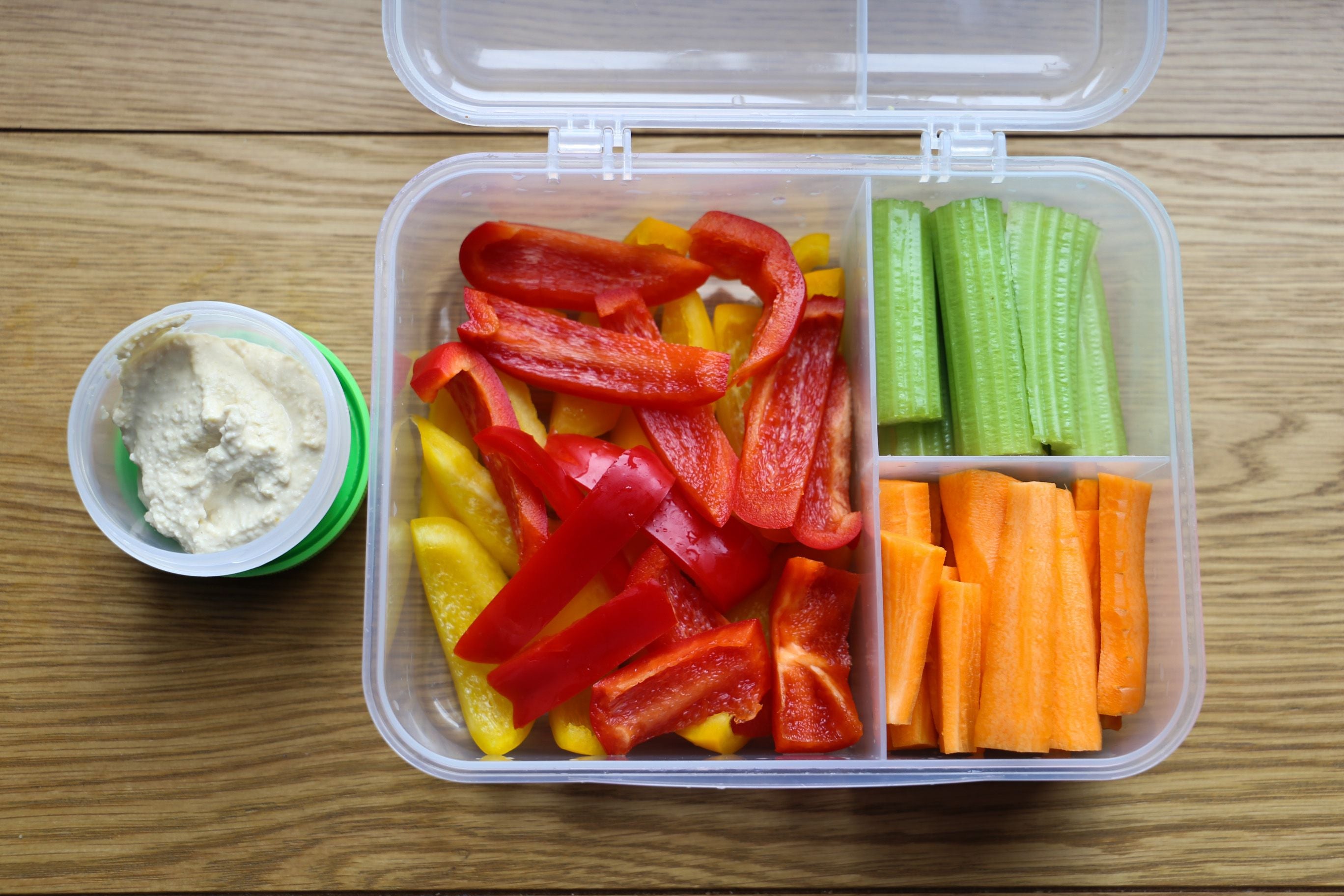 Brotdose mit Hummus, Sellerie-, Karotten- und Paprikasticks auf Holztisch.