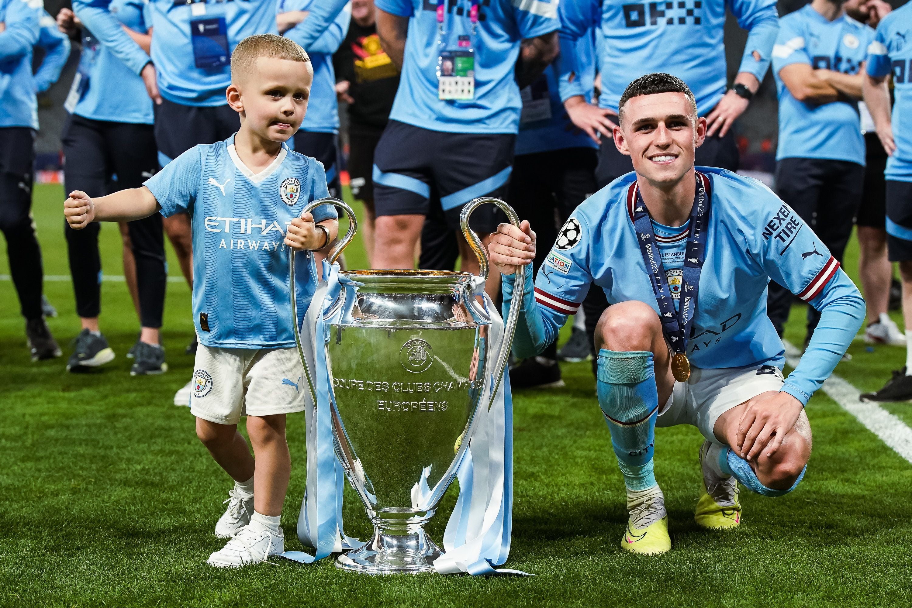 Ein Fußballspieler und ein Kind posieren mit einem großen Pokal auf einem Fußballfeld.