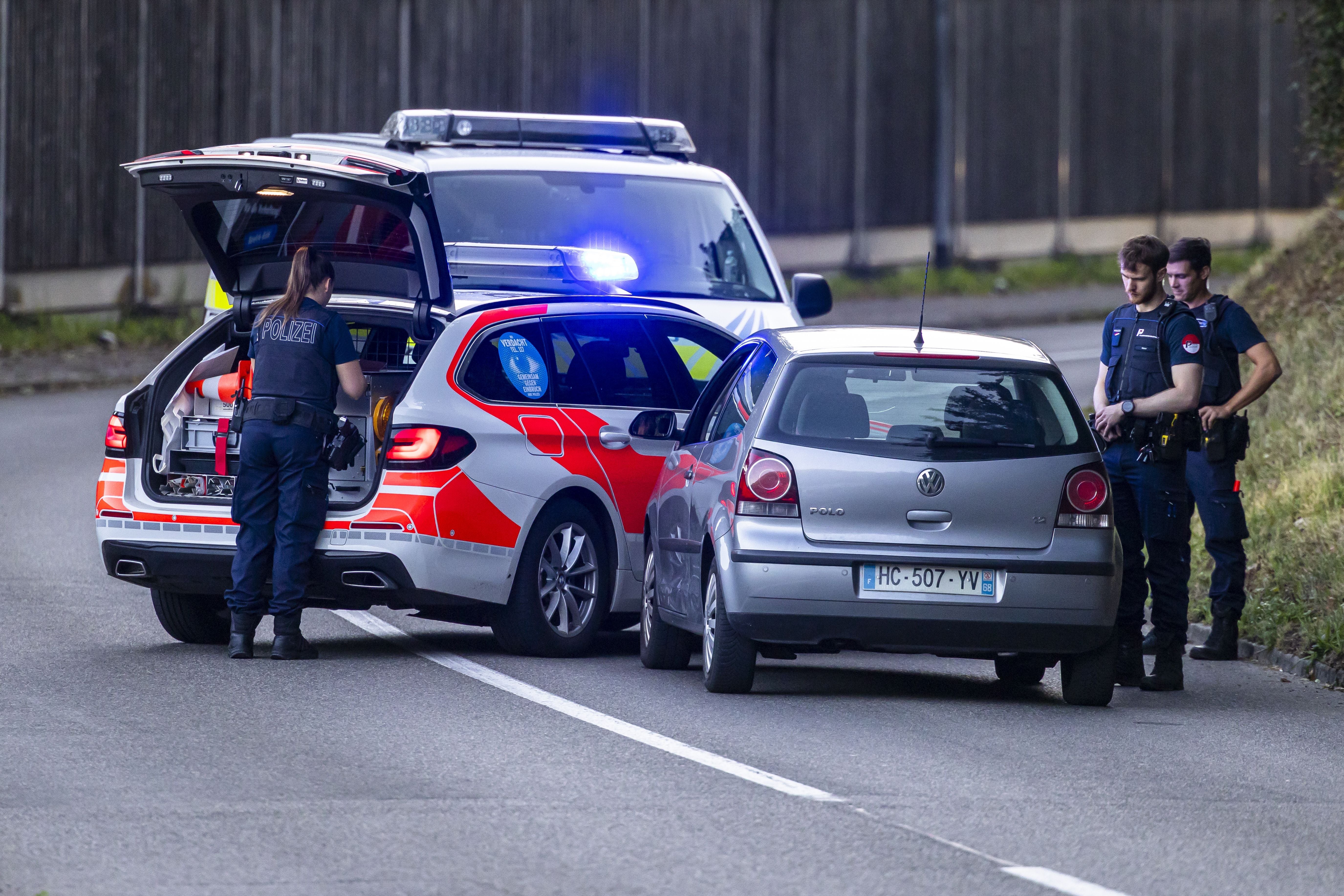 Polizisten kontrollieren Autos und blockieren eine Straße mit Einsatzfahrzeug und Blaulicht.