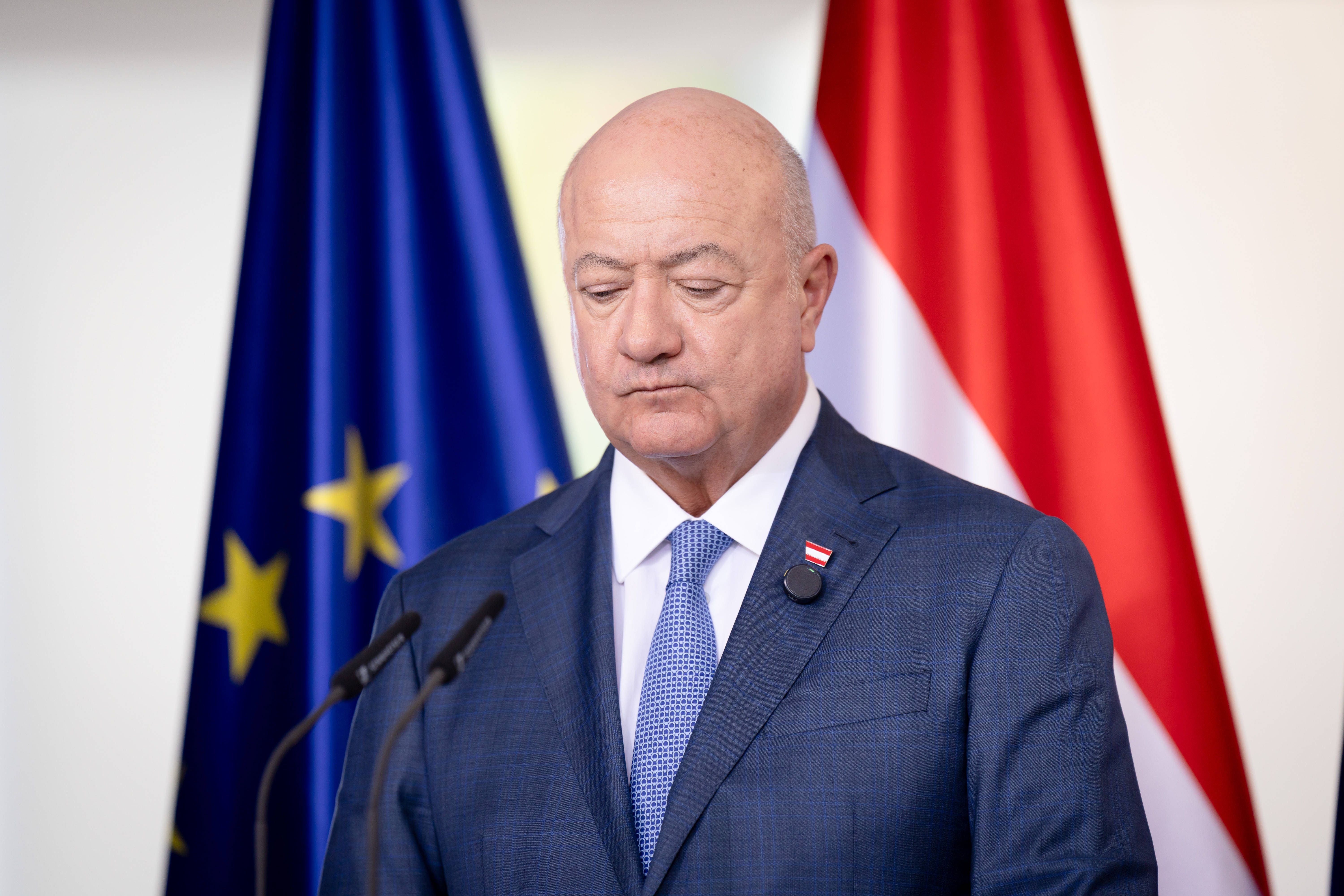 Ein Mann im Anzug steht nachdenklich vor Mikrofonen, im Hintergrund EU- und Österreich-Flagge.