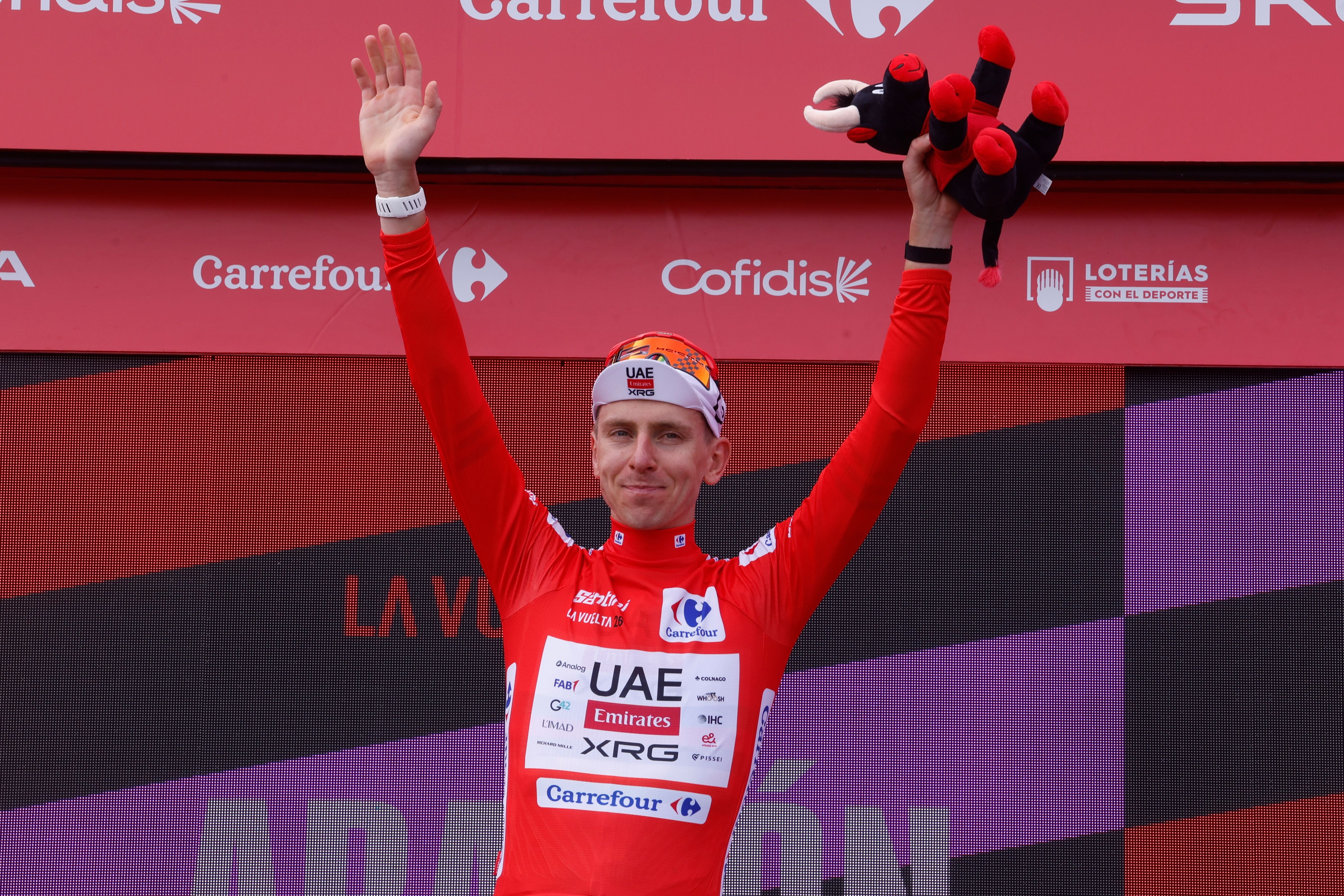 Ein Radrennfahrer im roten Trikot jubelt auf dem Podium und hält ein Stofftier in die Höhe.