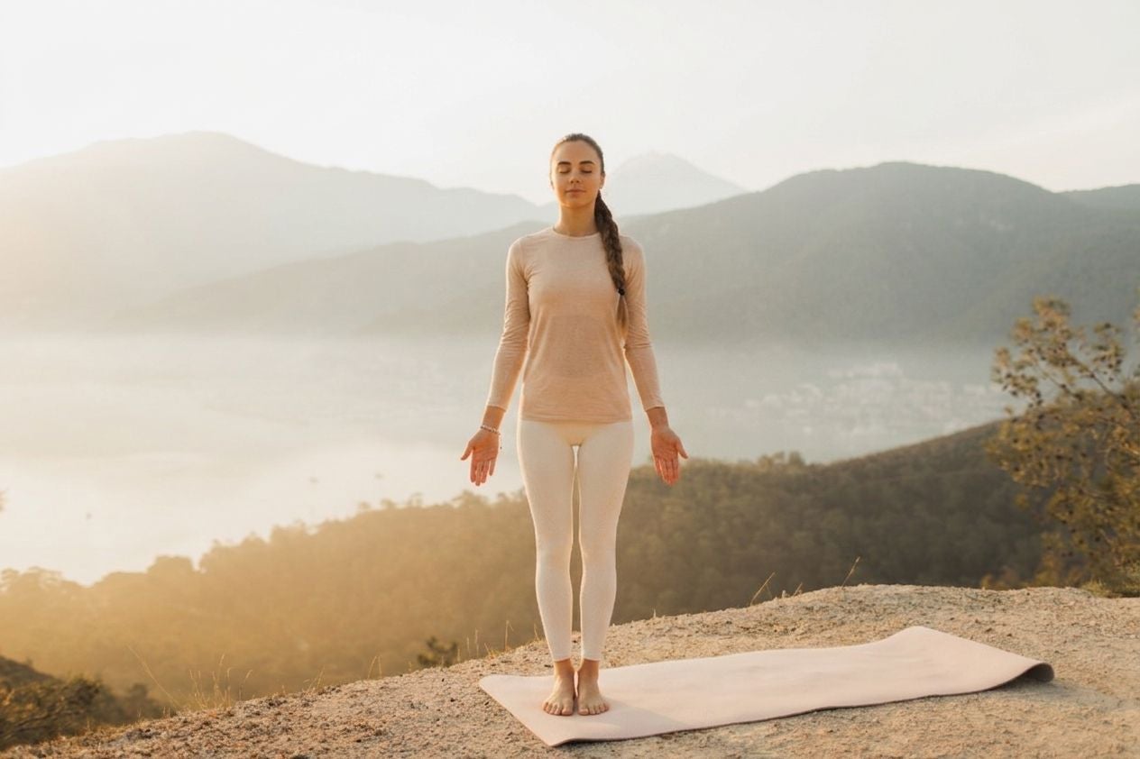Frau meditiert im Stehen auf einer Yogamatte in der Natur mit Bergen im Hintergrund.