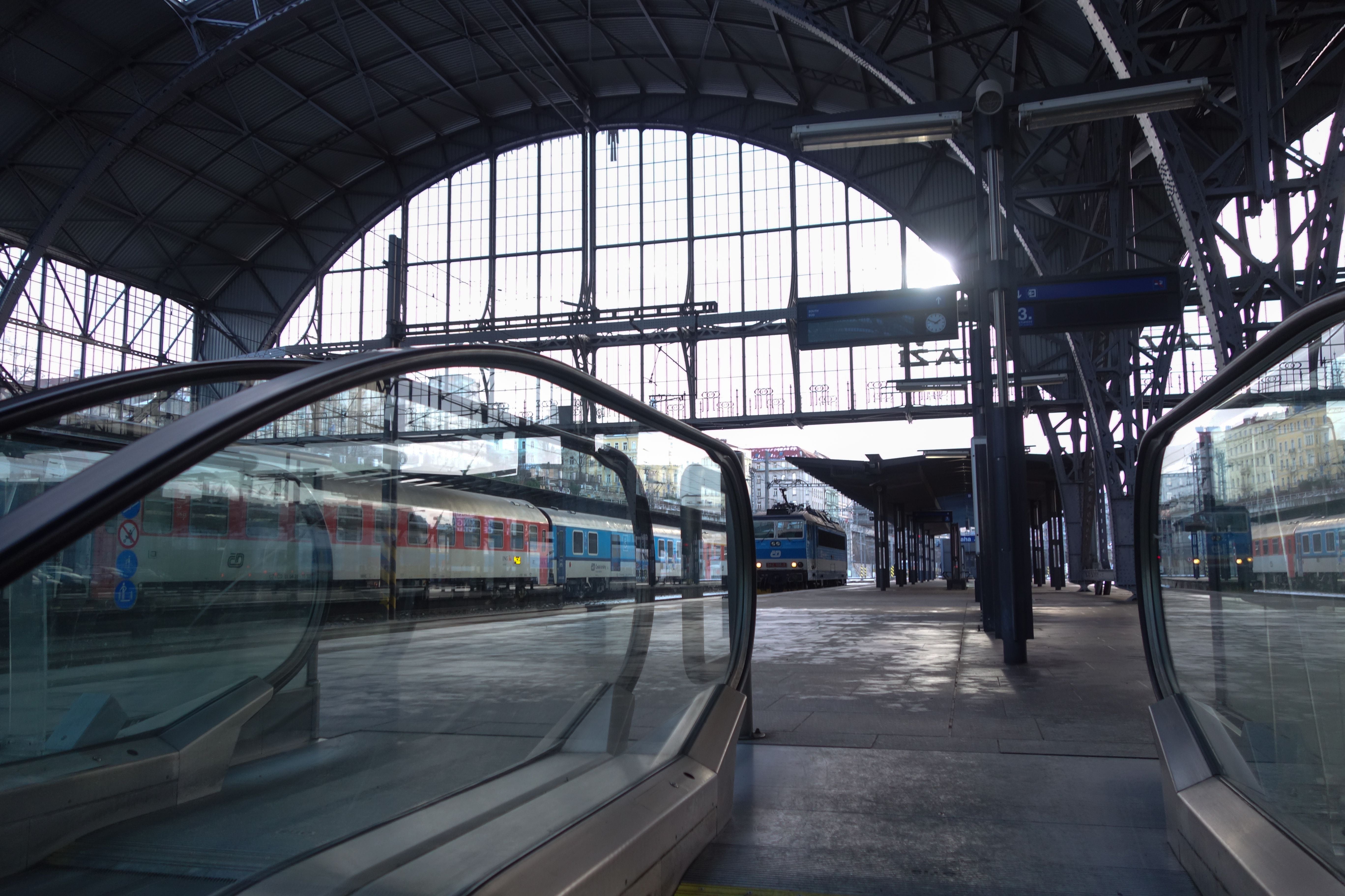 Blick auf einen Bahnsteig mit Rolltreppe im Hauptbahnhof Prag und Zügen im Hintergrund.
