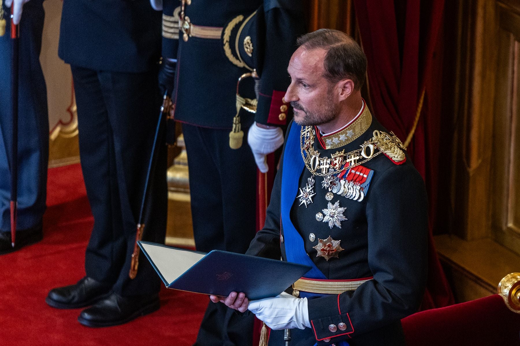 Ein Mann in königlicher Uniform mit Ordensschmuck hält ein offenes Buch bei einer Zeremonie.