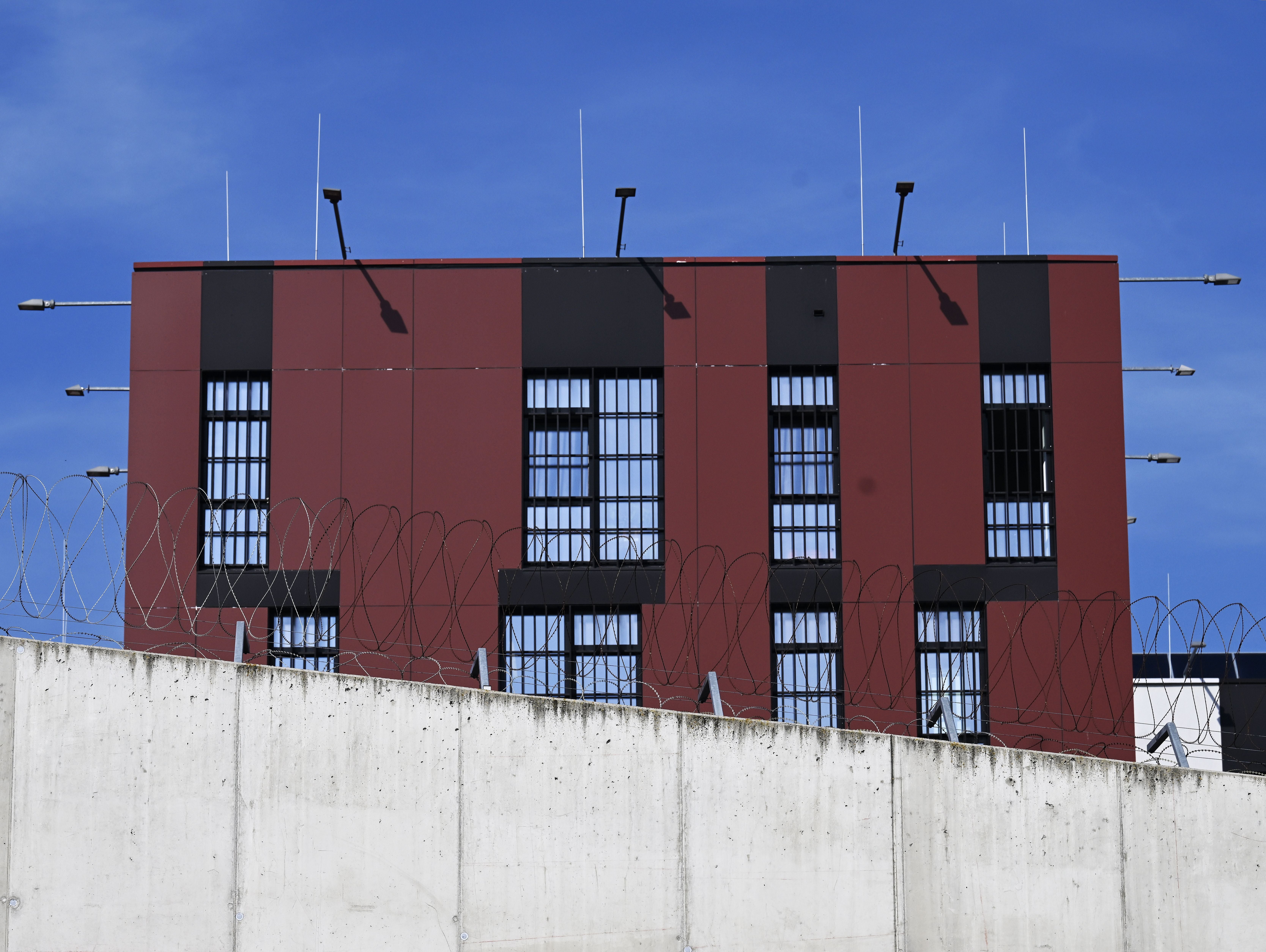 Gefängnis mit vergitterten Fenstern, Stacheldraht und hoher Mauer vor blauem Himmel.