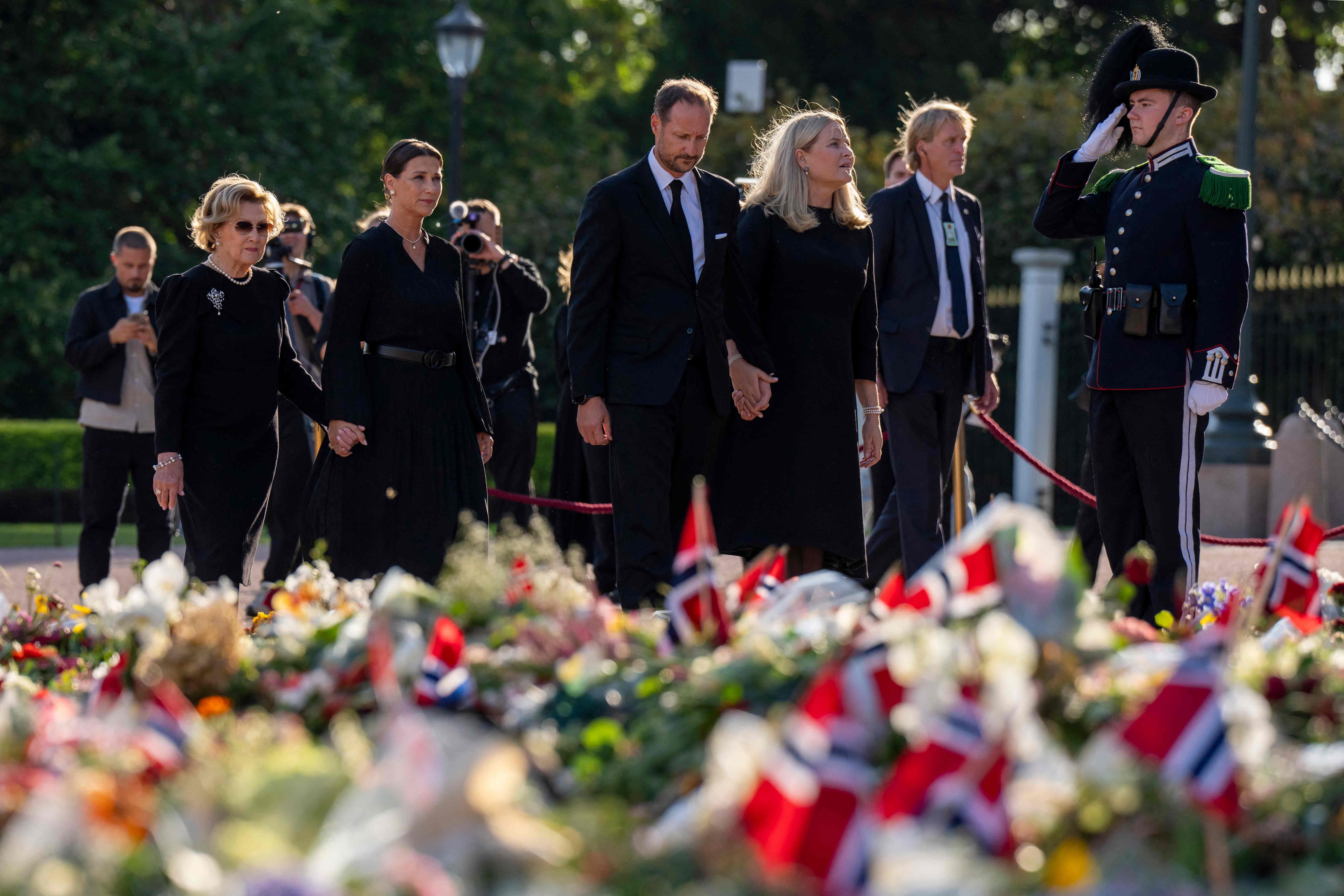 Mehrere Personen in schwarzer Kleidung stehen vor Blumen und norwegischen Fahnen in Gedenken.