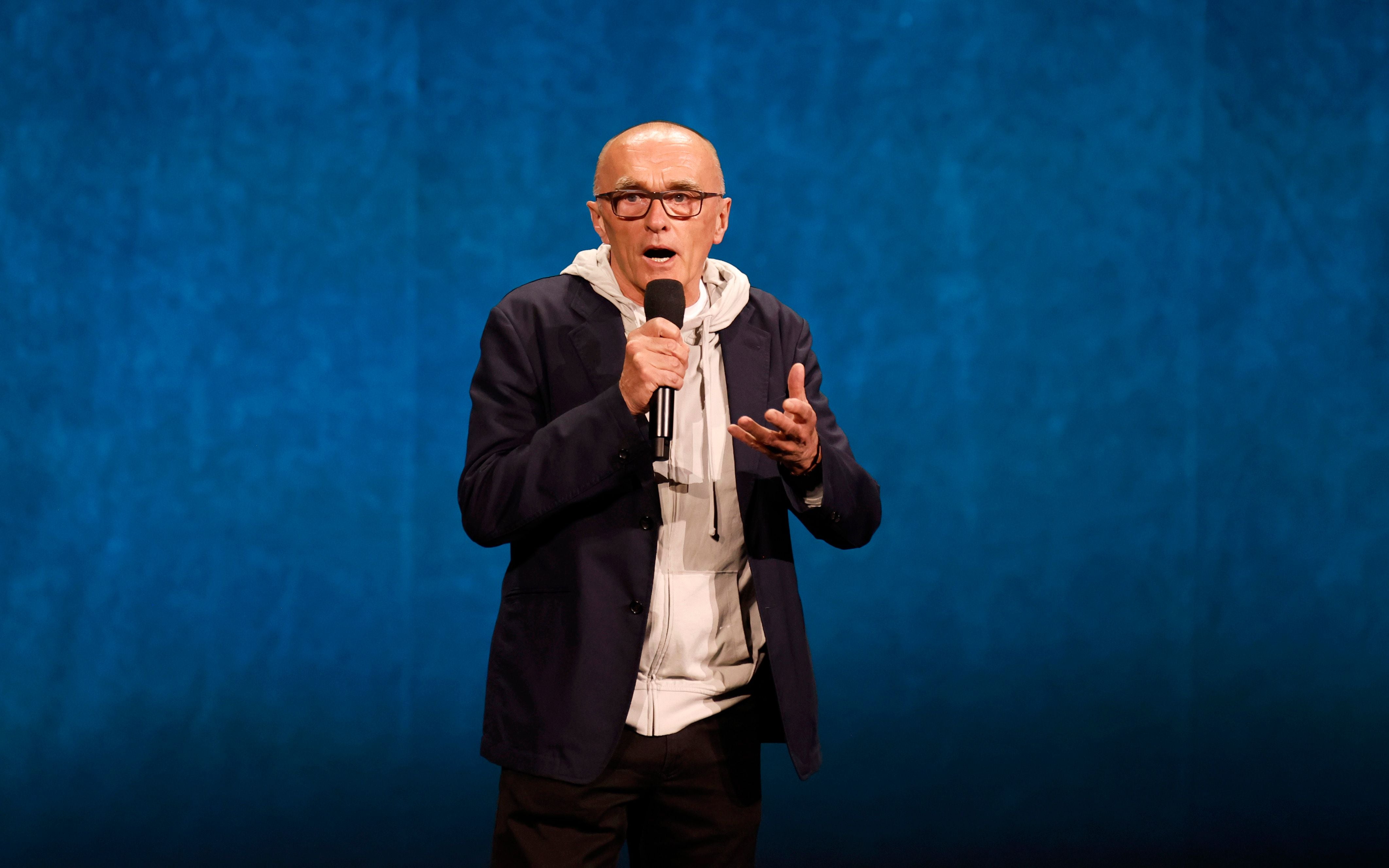 Ein Mann mit Brille spricht auf einer Bühne und hält ein Mikrofon vor blauem Hintergrund.