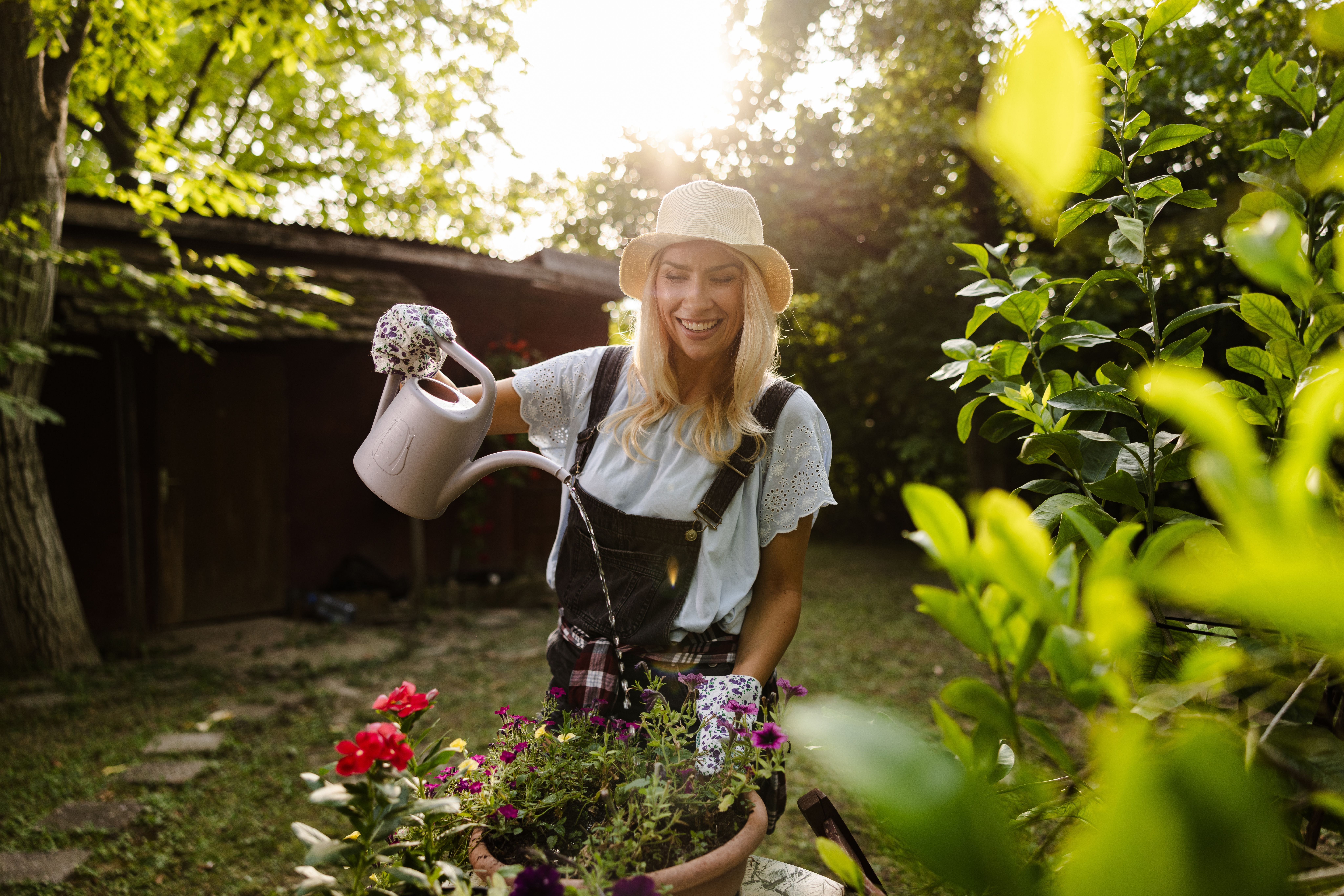 Fröhliche Frau gießt Blumen im Garten bei Sonnenlicht, umgeben von viel grünem Laub.