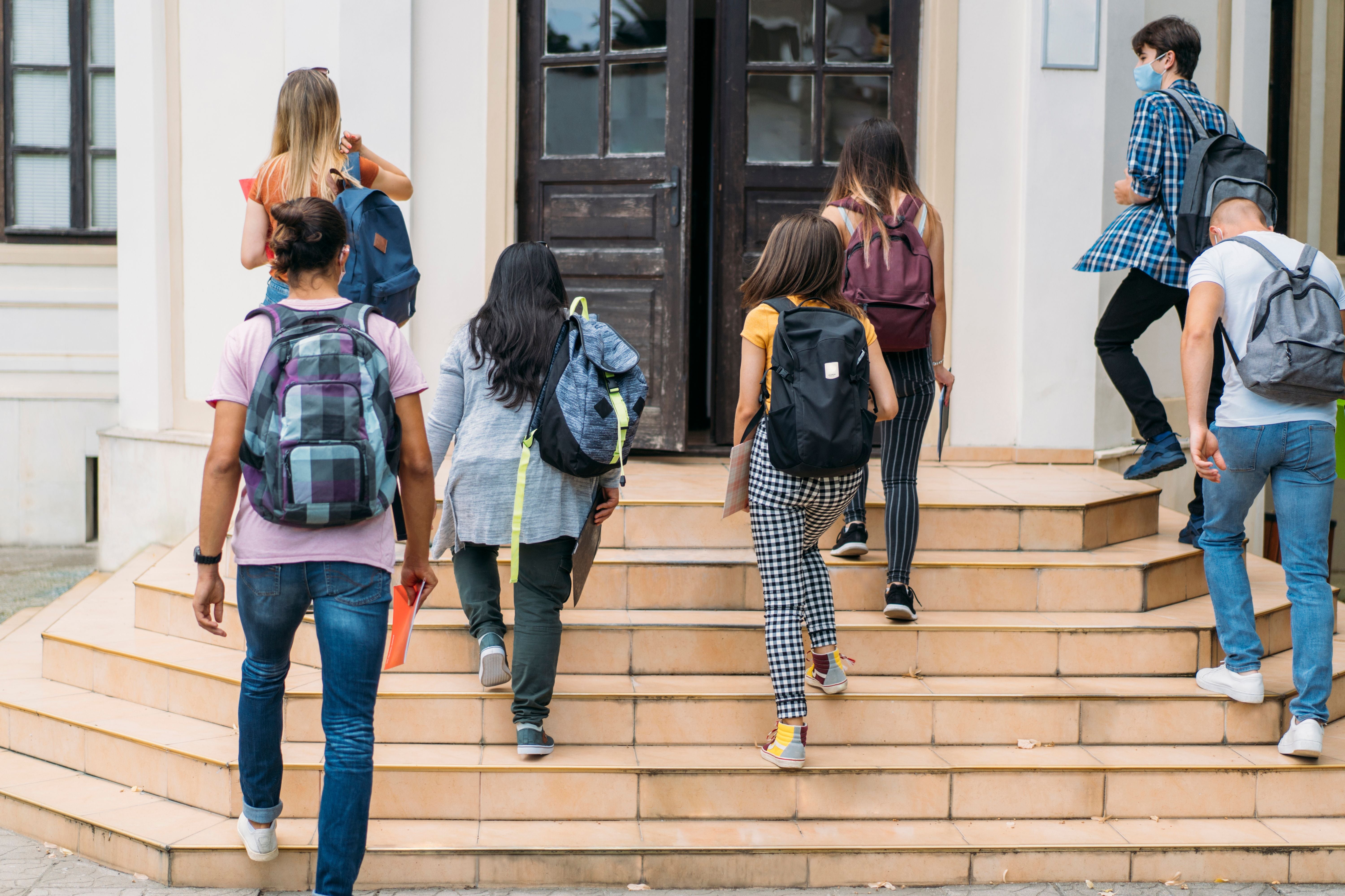 Mehrere Schüler mit Rucksäcken gehen eine Treppe zu einem Schulgebäude hinauf, einige tragen Masken.