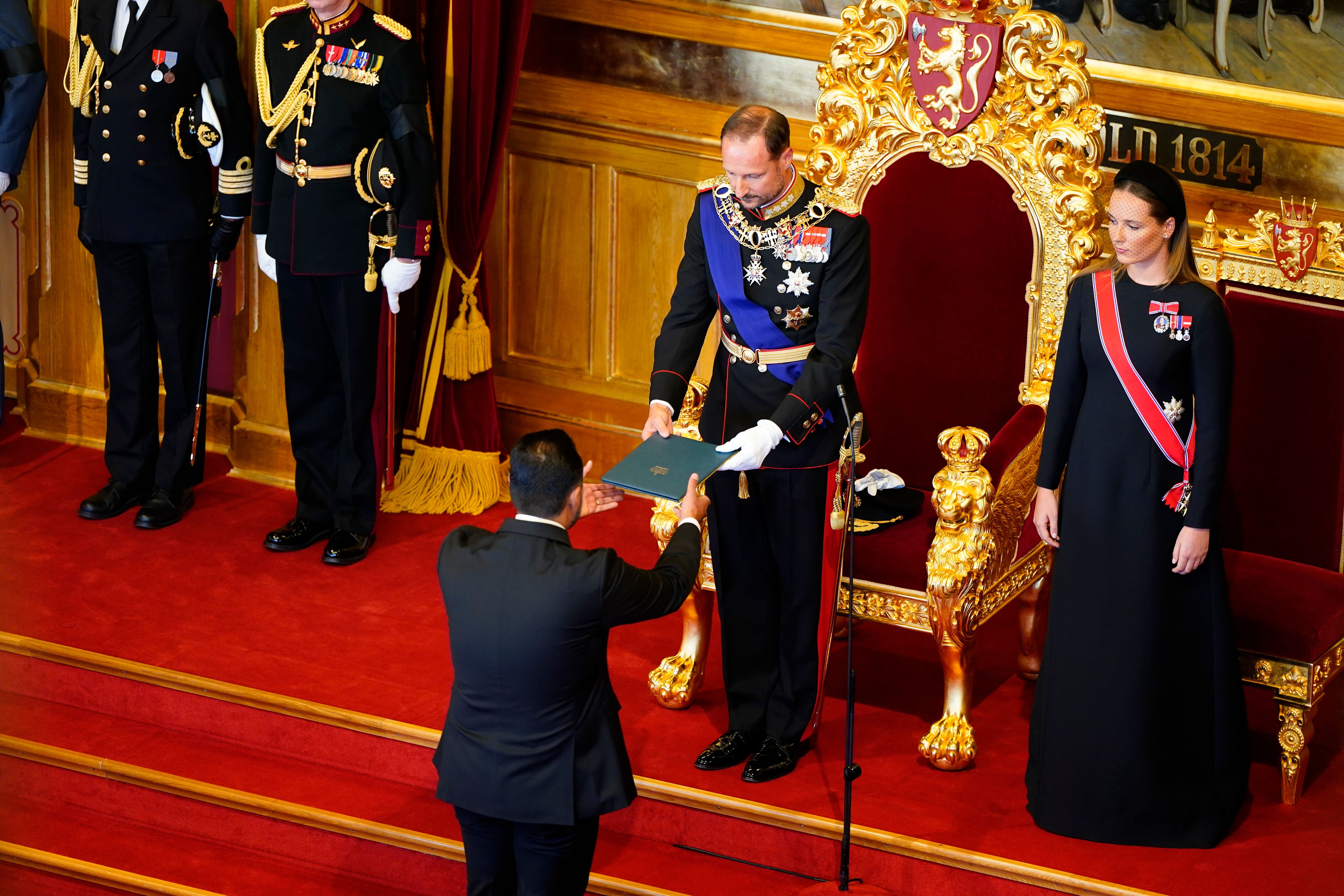 Ein Mann überreicht dem norwegischen König in Uniform ein Dokument im Parlament auf rotem Teppich.