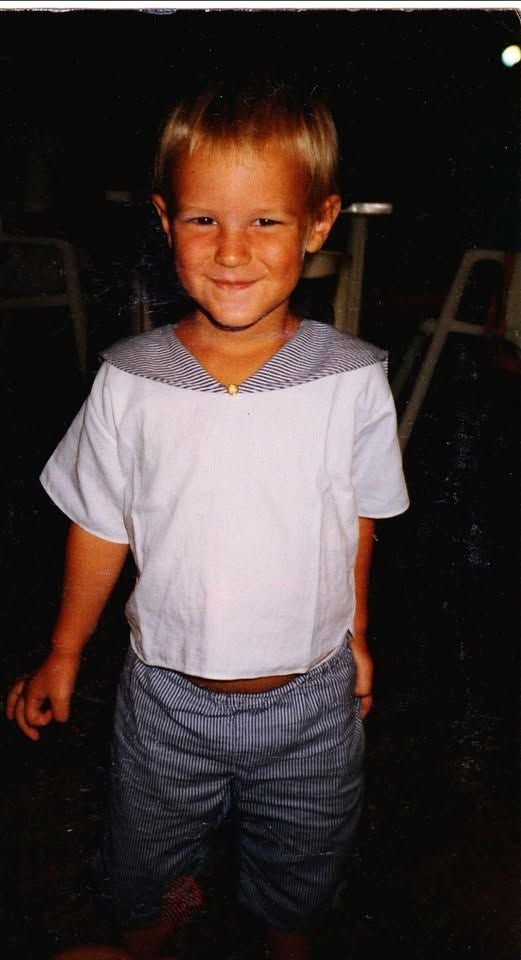 Ein Kind mit kurzem blondem Haar lächelt und trägt ein weißes Matrosenshirt und gestreifte Shorts.