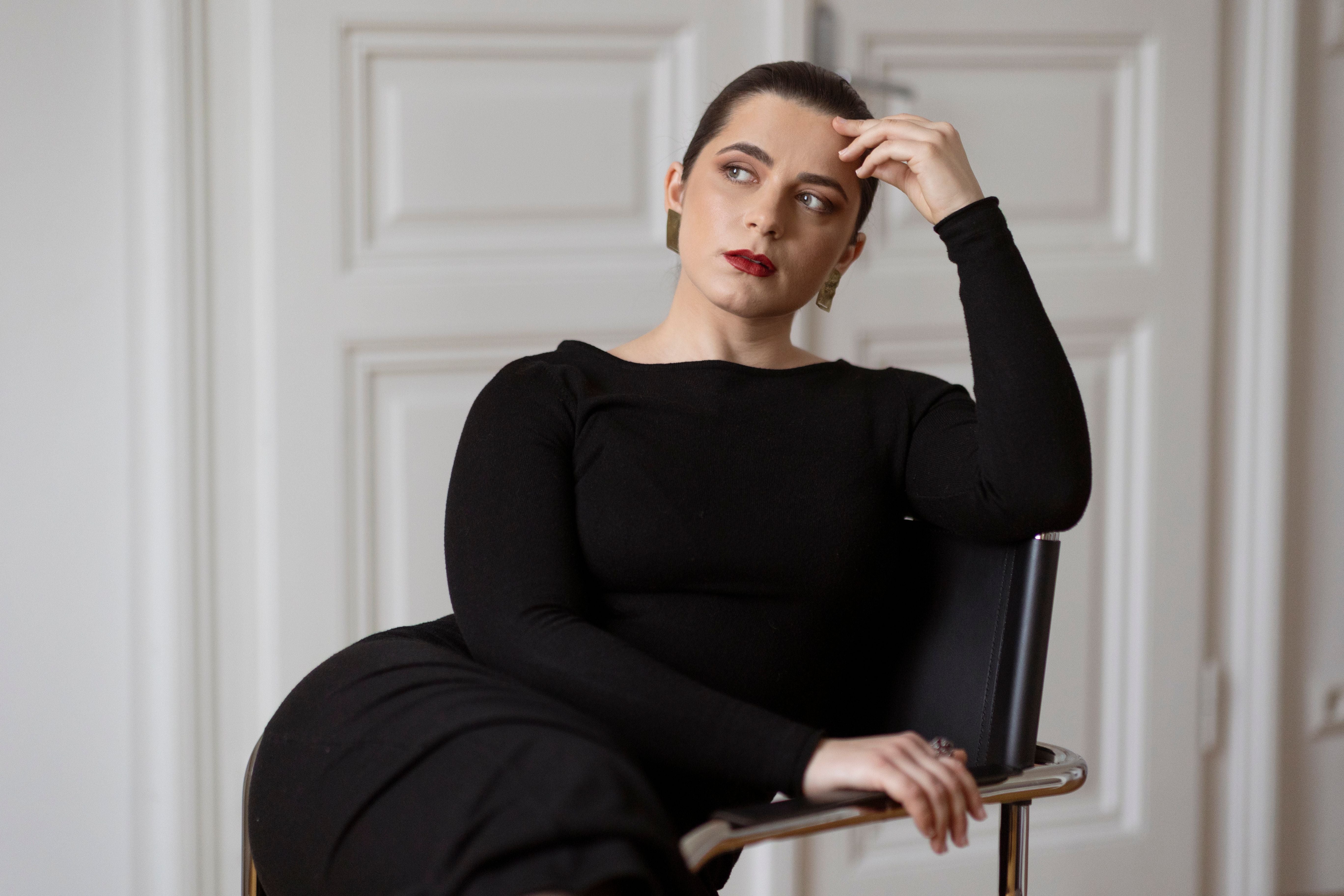 Frau in schwarzem Kleid sitzt auf einem Stuhl und schaut nachdenklich zur Seite.