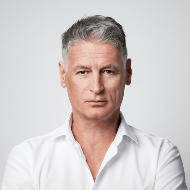 Ein Mann mit grauen Haaren trägt ein weißes Hemd und blickt ernst in die Kamera.