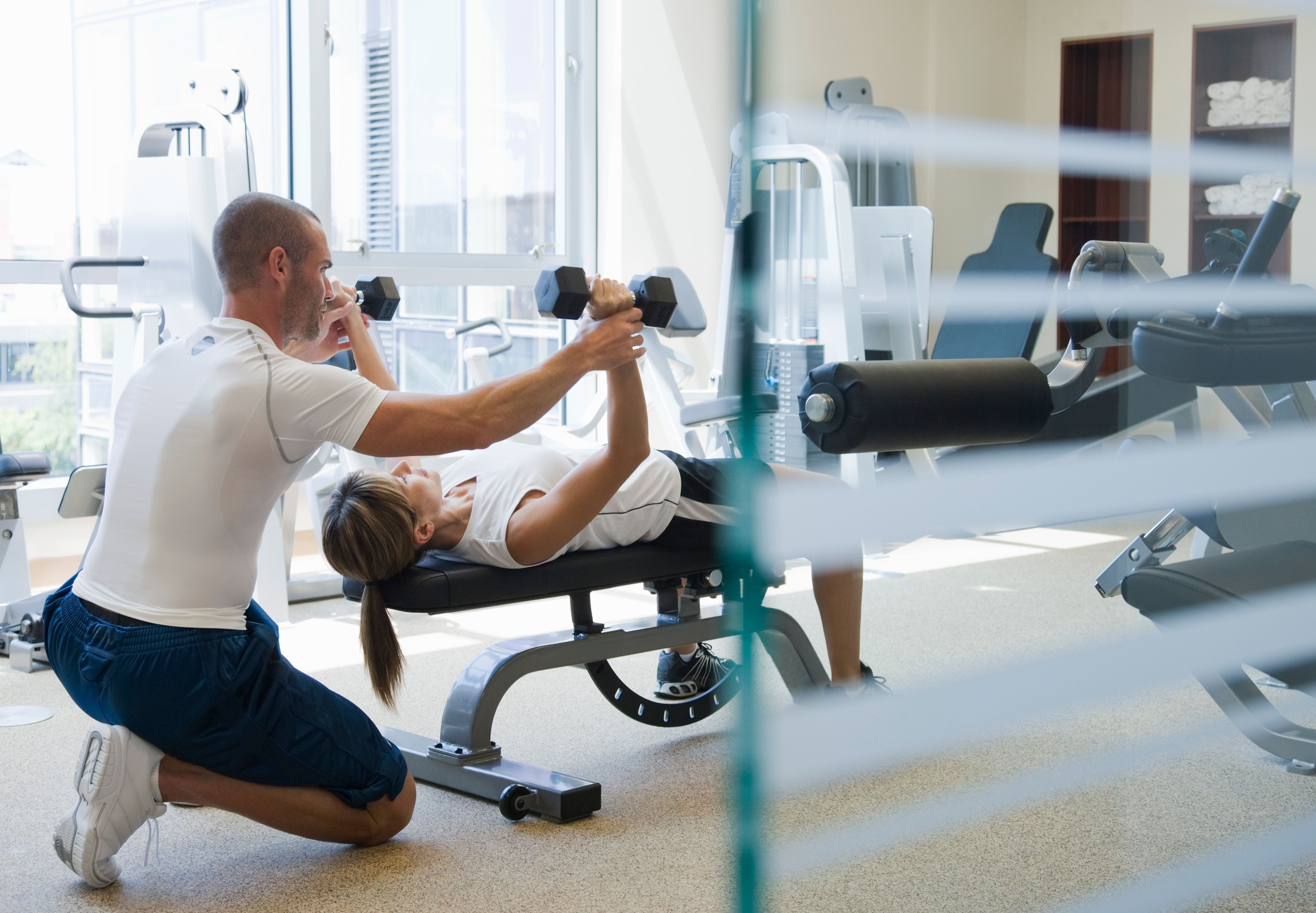 Trainer unterstützt Frau beim Bankdrücken mit Kurzhanteln im Fitnessstudio.