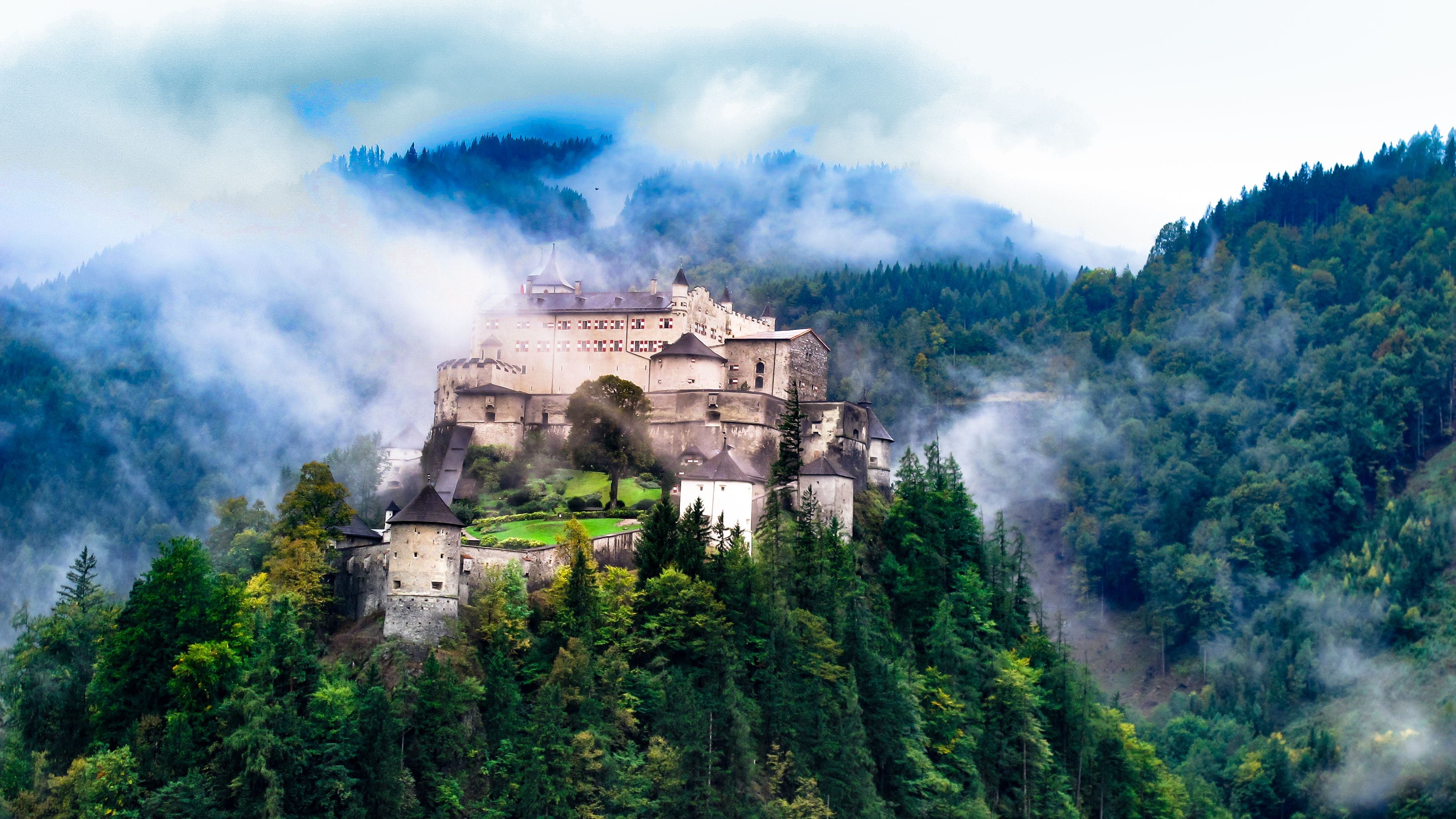 Burg auf bewaldetem Hügel, umgeben von Nebel und dichten Bäumen in bergiger Landschaft.