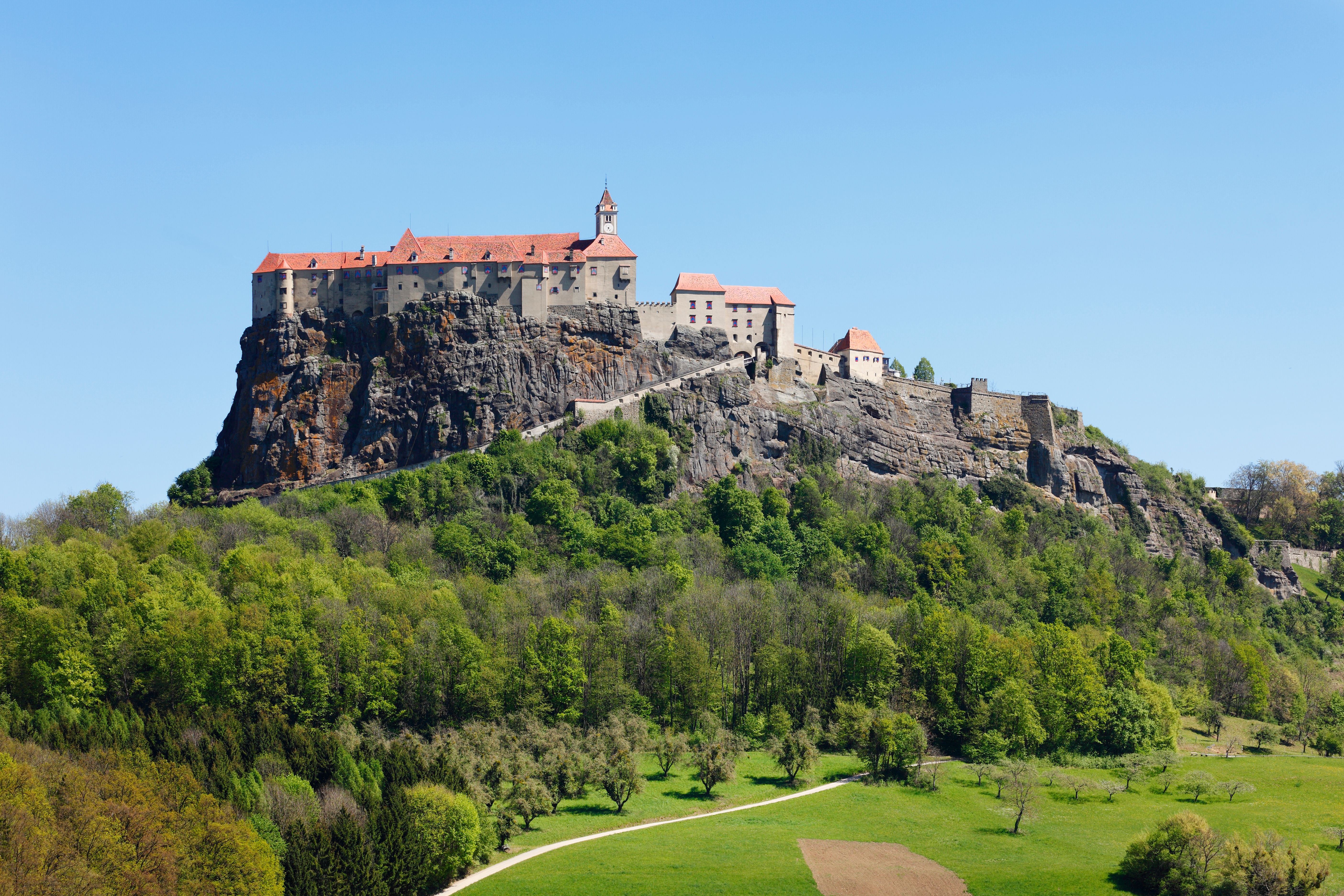 Burg mit rotem Dach thront auf einem Felsen, umgeben von grünen Wäldern und Wiesen.