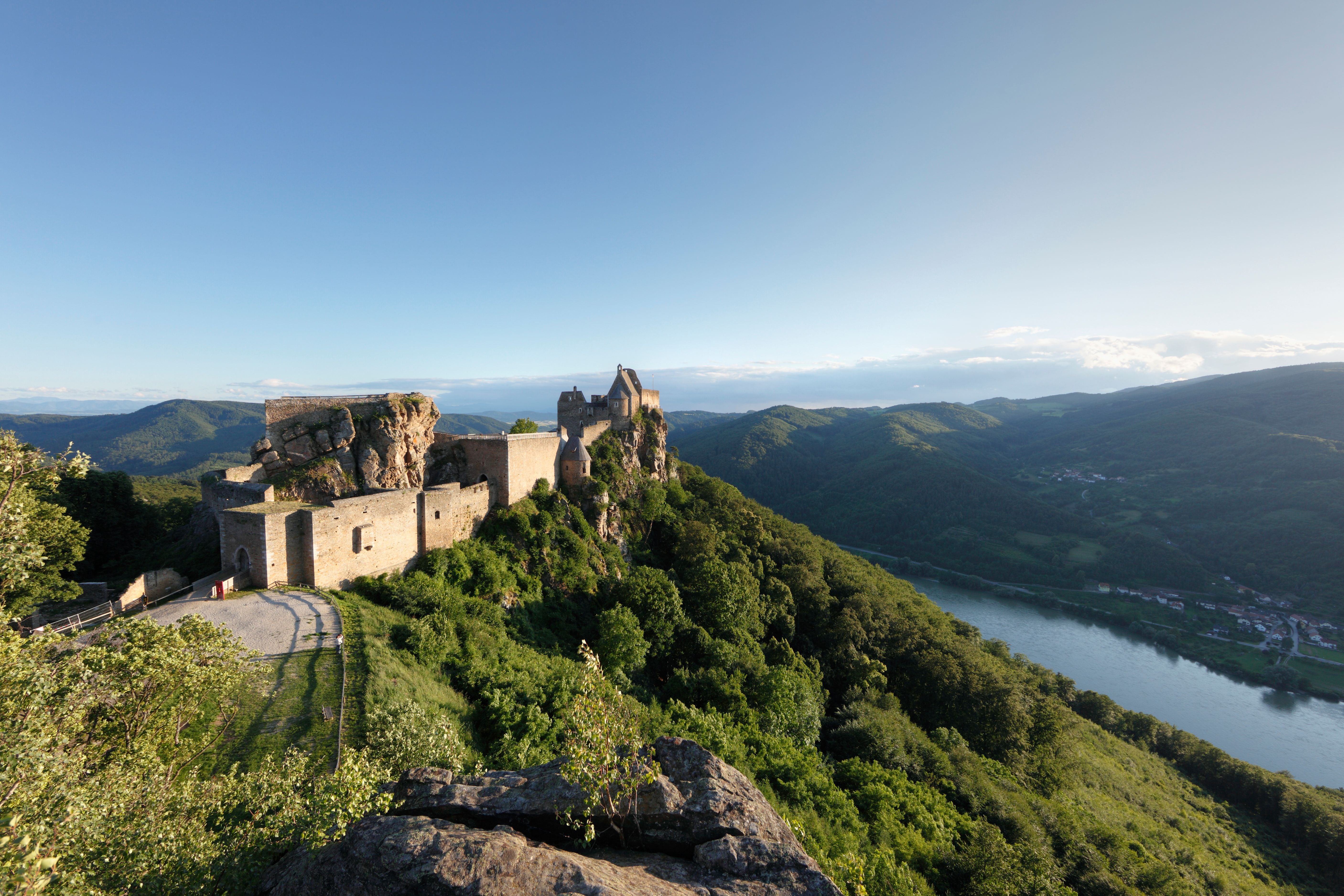 Burgruine auf einem Hügel mit Blick auf bewaldete Berge und einen Fluss im Tal.