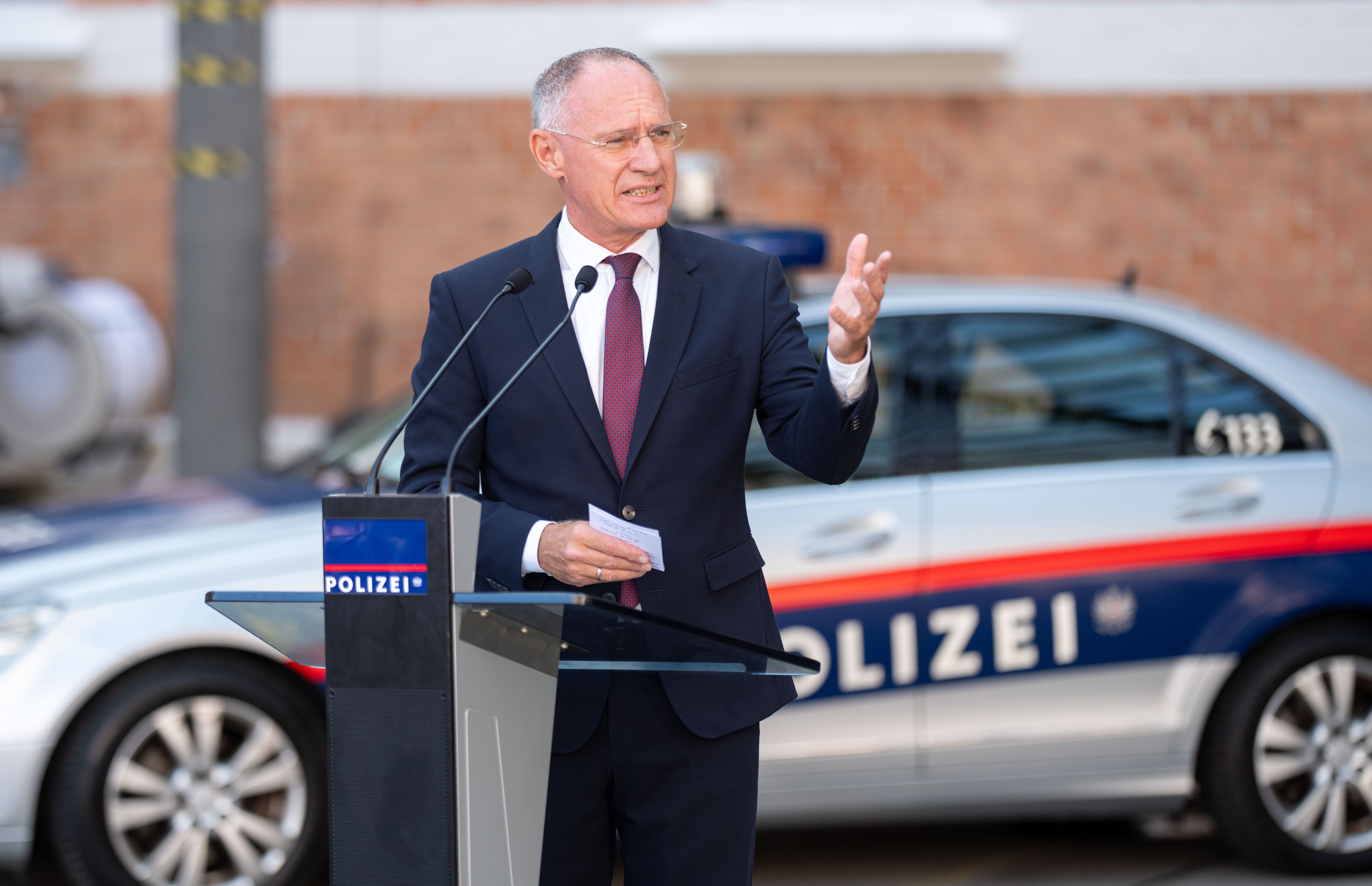 Ein Mann im Anzug spricht bei einer Polizei-Veranstaltung vor einem Polizeiauto und Mikrofon.