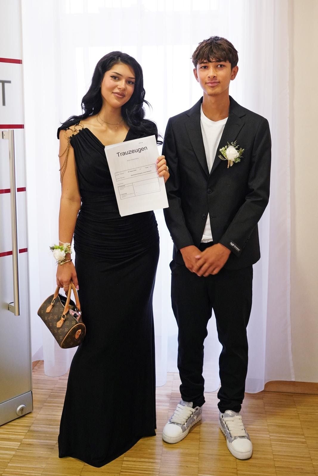 Eine Frau im schwarzen Kleid und ein Mann im Anzug stehen mit Trauzeugen-Formular nebeneinander.