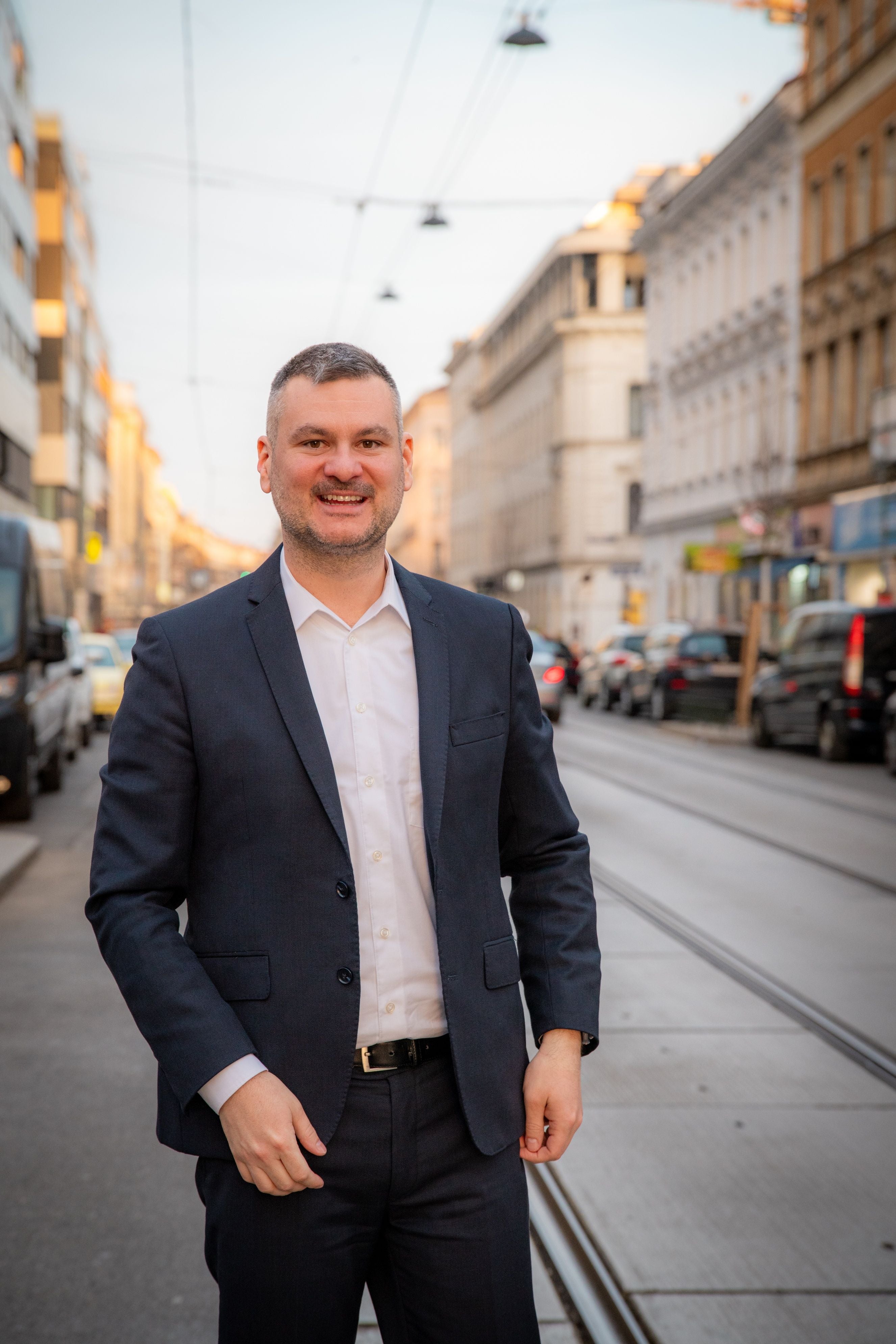 Mann im Anzug steht lächelnd auf einer städtischen Straße mit Häusern im Hintergrund.