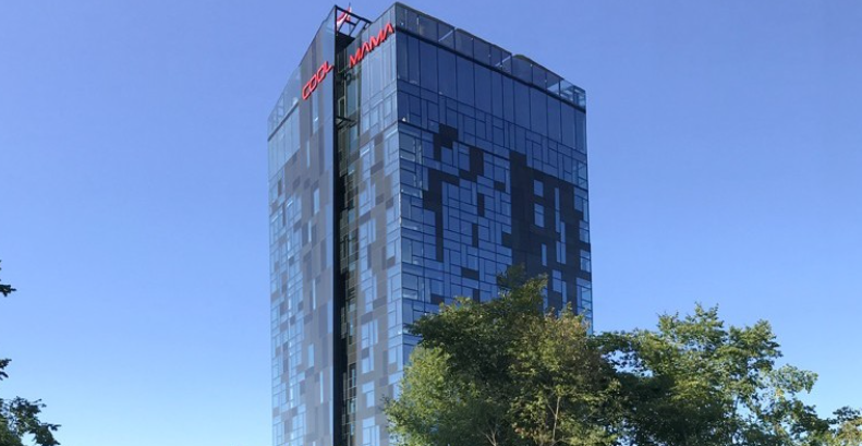 Modernes Hochhaus mit Glasfassade und rotem Schriftzug "COOL MAMA" oben, Bäume im Vordergrund.