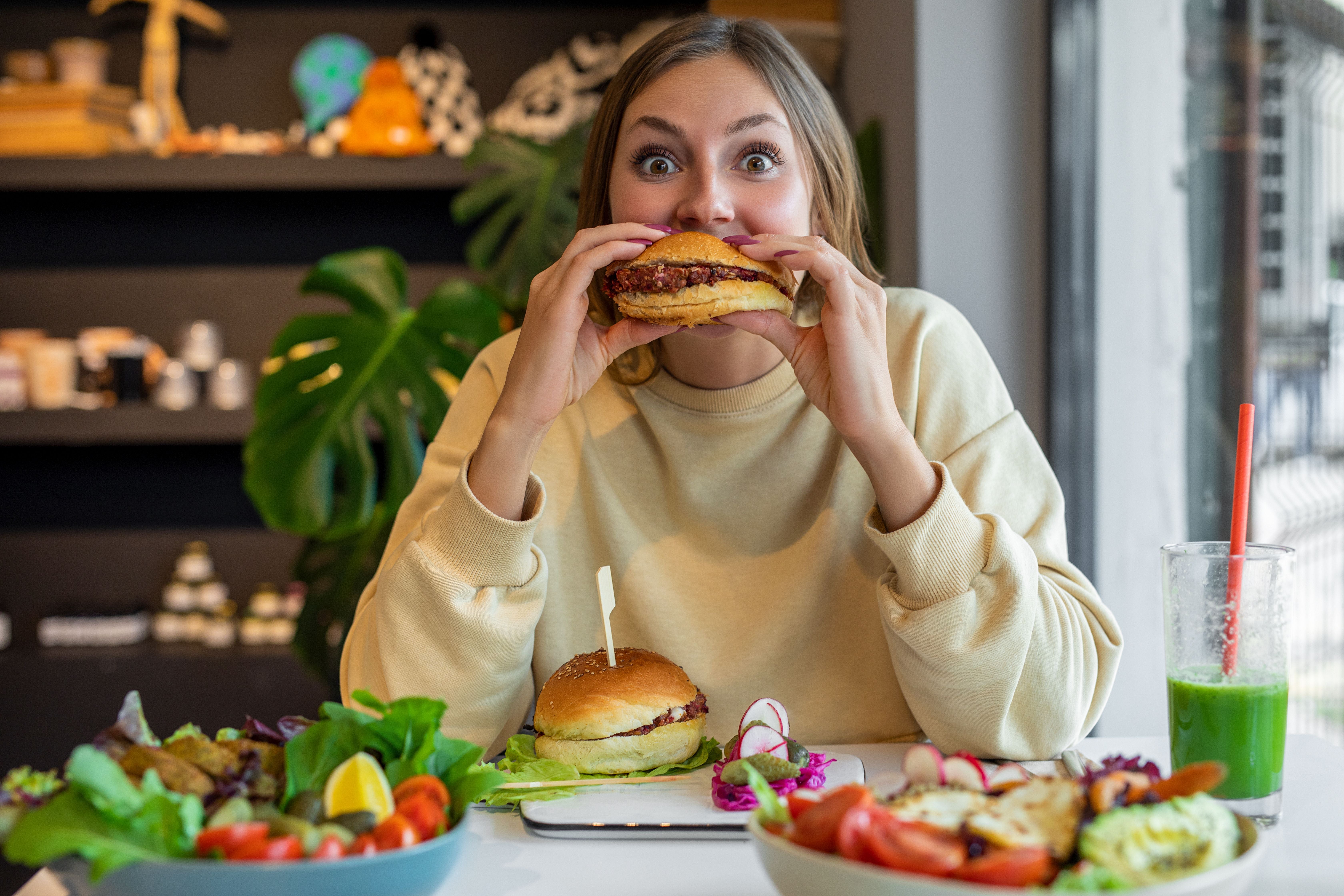 Frau isst einen veganen Burger in einem Café, mit Salat und grünem Saft auf dem Tisch.