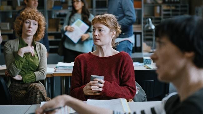 Drei Frauen sitzen in einem Raum, eine davon hält eine Tasse, im Hintergrund weitere Personen.